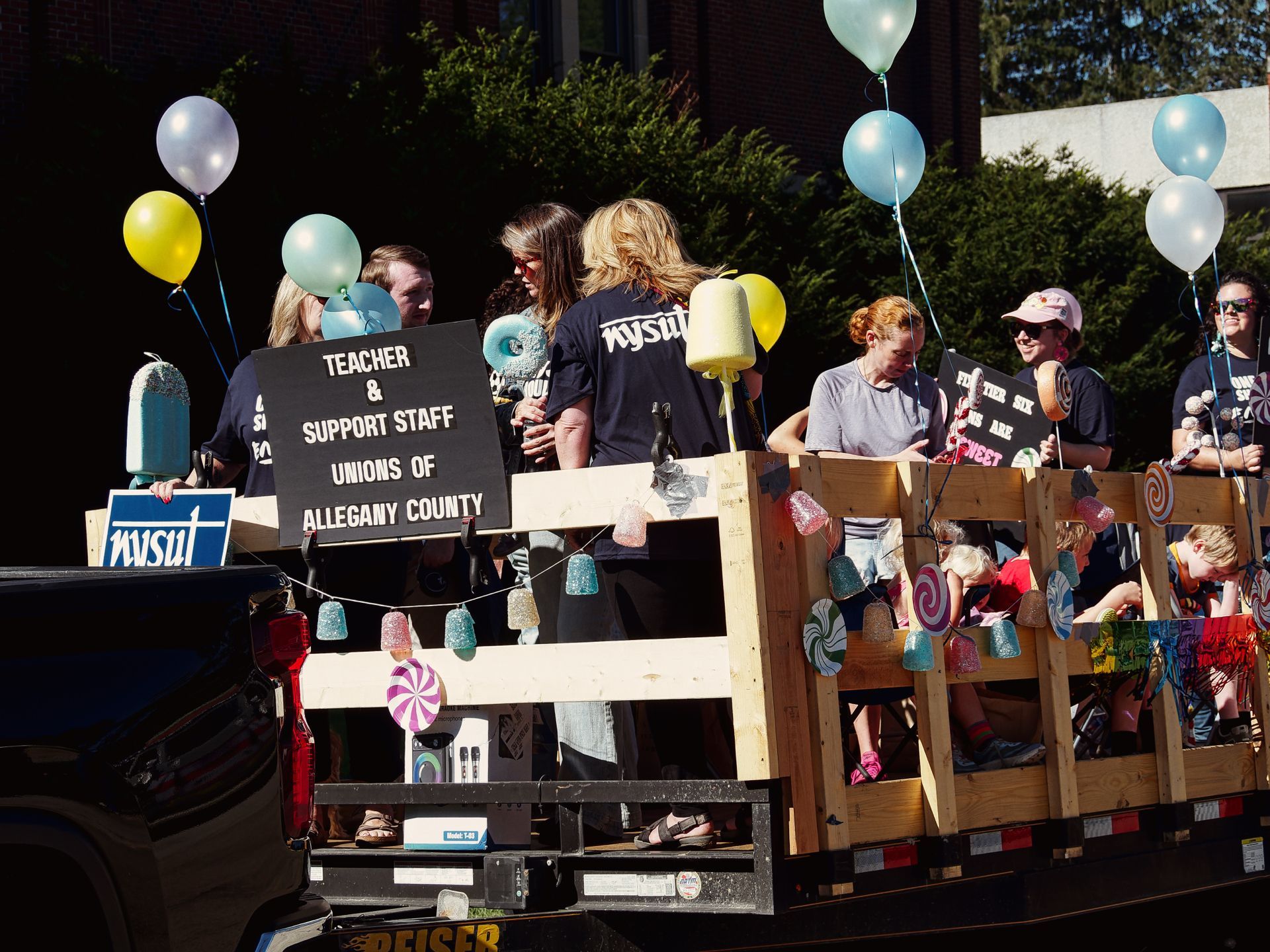 Parade float pulled by a dark pickup truck, decorated with candy-themed ornaments — glittery bells, pinwheel lollipops, and oversized lollipop props — and clusters of white, yellow, and blue balloons. Adults in navy "NYSUT" t-shirts stand on the wooden flatbed alongside young children; a black sign in the foreground reads "TEACHER & SUPPORT STAFF UNIONS OF ALLEGANY COUNTY," and a blue "NYSUT" placard is visible on the left rail. A brick building and dense trees sit in the shadowed background.