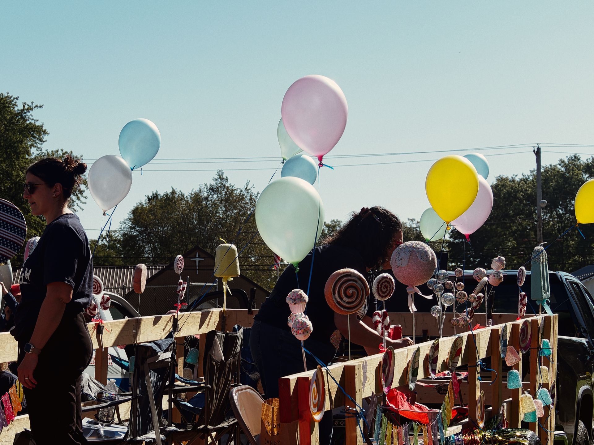 Two women in black shirts work alongside a wooden parade float decorated with oversized lollipop props and clusters of pastel balloons — pink, mint, white, blue, and yellow — floating against a clear midday sky, with trees, rooftops, and a utility pole visible in the background.