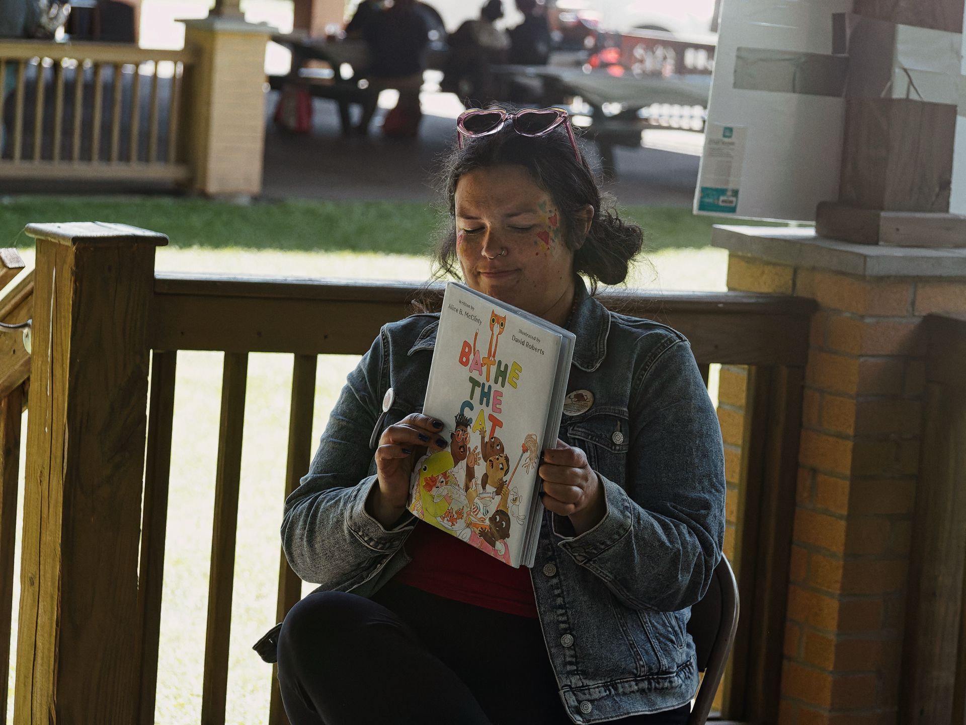 Woman with heart-shaped sunglasses pushed up on her head and colorful face paint dots on her cheeks, seated on a wooden deck, holding open the children's book *Bathe the Cat* — illustrated by David Roberts — toward the camera. A grassy area and blurred picnic tables fill the background.