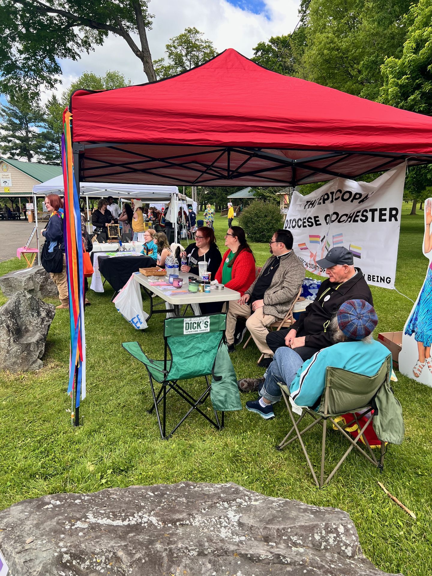 Several people seated in folding camp chairs — including a green Dick's Sporting Goods chair in the foreground — beneath a red pop-up canopy at an outdoor fair, with a banner reading "THE EPISCOPAL DIOCESE ROCHESTER" and partial text about dignity and being visible behind them. A rainbow ribbon streamer hangs from the canopy's left pole, and a cardboard cutout of a figure in a blue dress stands at the far right; additional vendor tents and green trees fill the background.