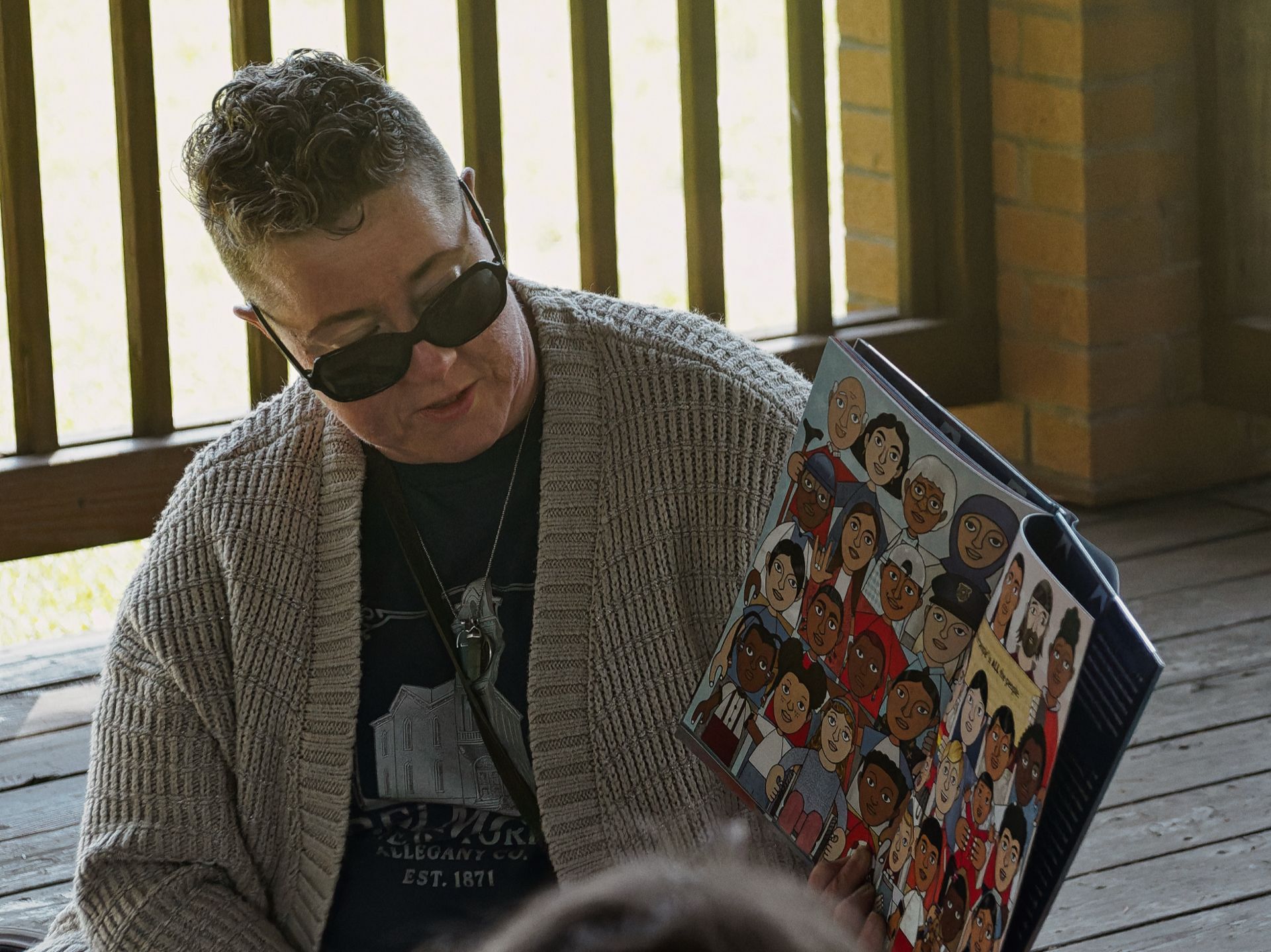 Person with short curly gray hair and dark sunglasses, wearing a beige knit cardigan over a black graphic tee reading "EST. 1871," seated on a wooden porch and looking down at a colorful illustrated children's book whose cover depicts a crowd of diverse cartoon characters.
