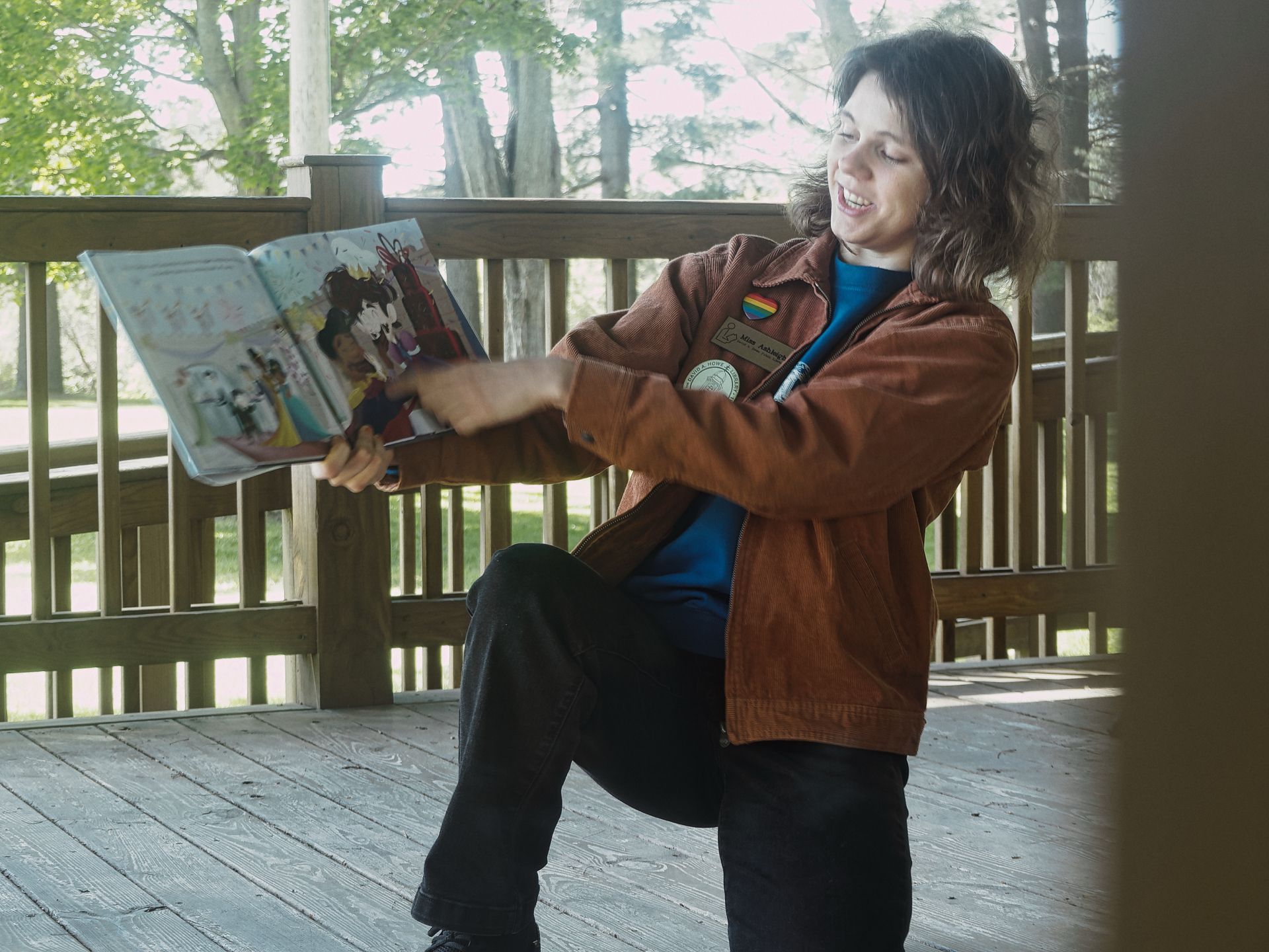 Person in a rust-brown corduroy jacket over a teal shirt, smiling and holding open a colorful illustrated children's book toward the camera on a wooden deck. A rainbow pride pin and a circular patch are visible on the jacket, with a pen tucked into the breast pocket. Trees and green lawn blur softly beyond the deck railing in the background.