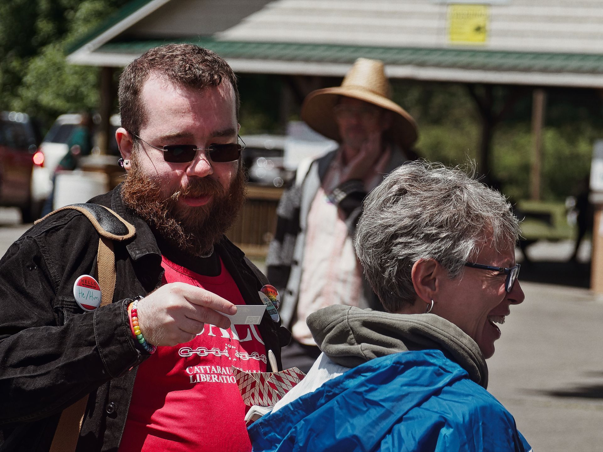 Bearded man in sunglasses, a red "Cattaraugus Liberation" t-shirt, black jacket, rainbow bracelet, and a "He/Him" name badge leans toward a laughing gray-haired woman in a blue jacket, holding out a small card or sticker; a third person in a straw hat stands blurred in the background beneath a covered outdoor structure.