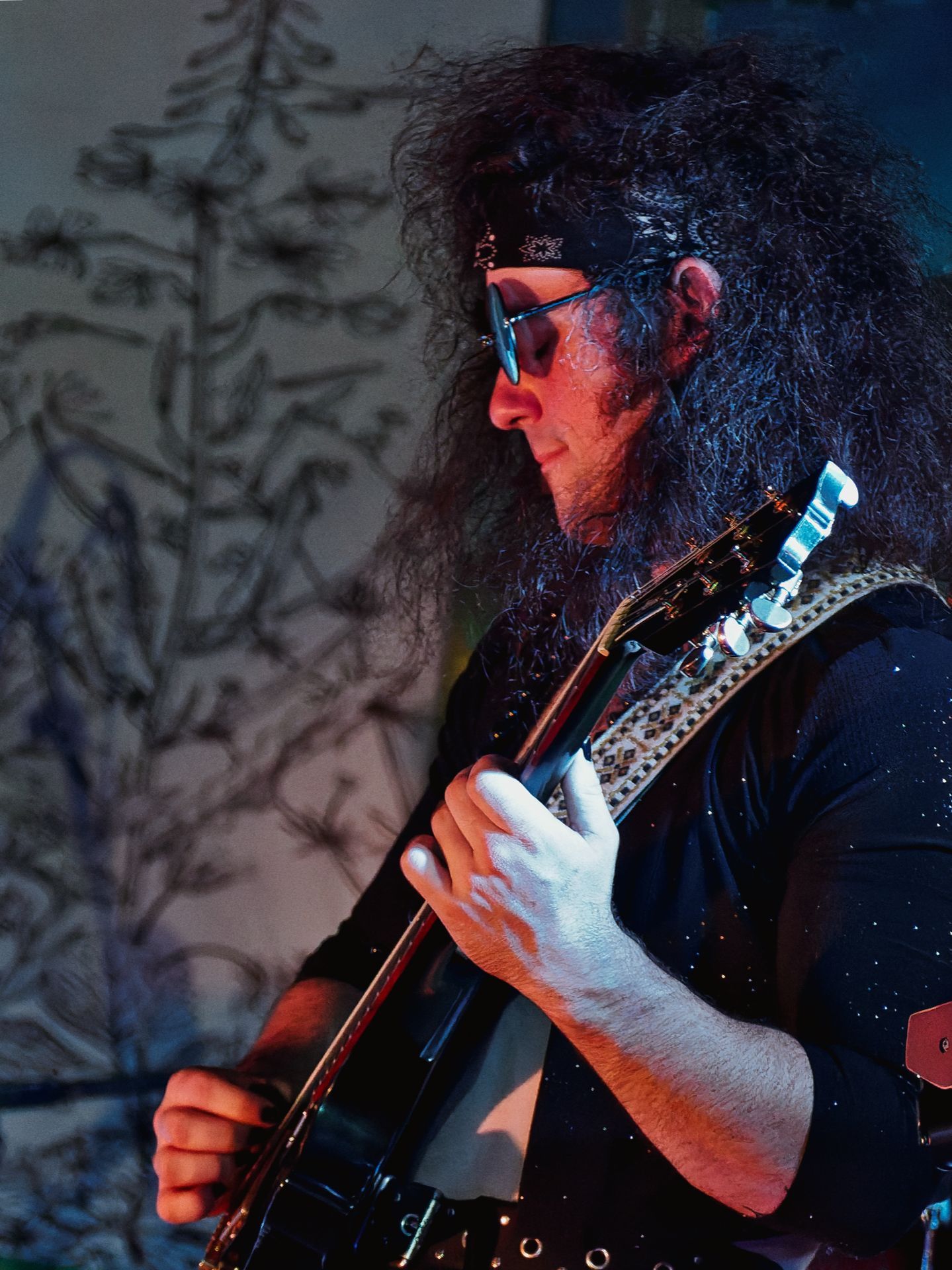 A guitarist with voluminous curly black hair, a dark bandana, and blue-tinted sunglasses plays a black electric guitar, lit from the left in cool blue and warm red tones. Behind him, a blurred wall mural of sketched botanical forms fades into the shallow background.
