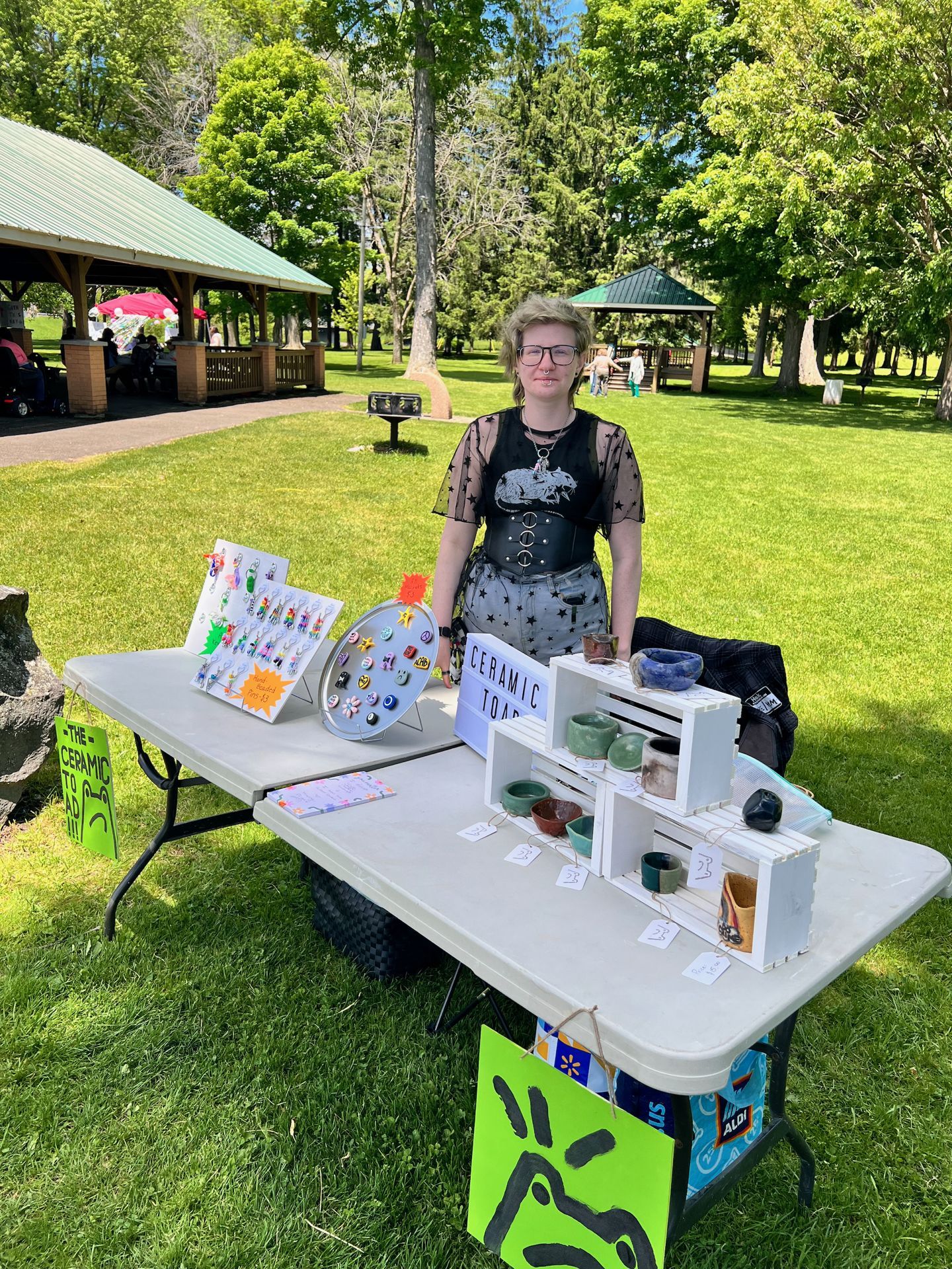 A young vendor with short blonde-green hair and glasses stands behind a folding table at an outdoor craft fair in a sunny park, wearing a sheer black star-print top and a wide leather corset belt. The table displays handmade ceramic bowls and cups arranged on white wooden crates, a circular pin display board with colorful enamel pins, and a colorful earring card, with a sign reading "CERAMIC TOAD" at center and a bright green sign to the left reading "THE CERAMIC TOAD" — a pavilion and tall trees visible in the background.