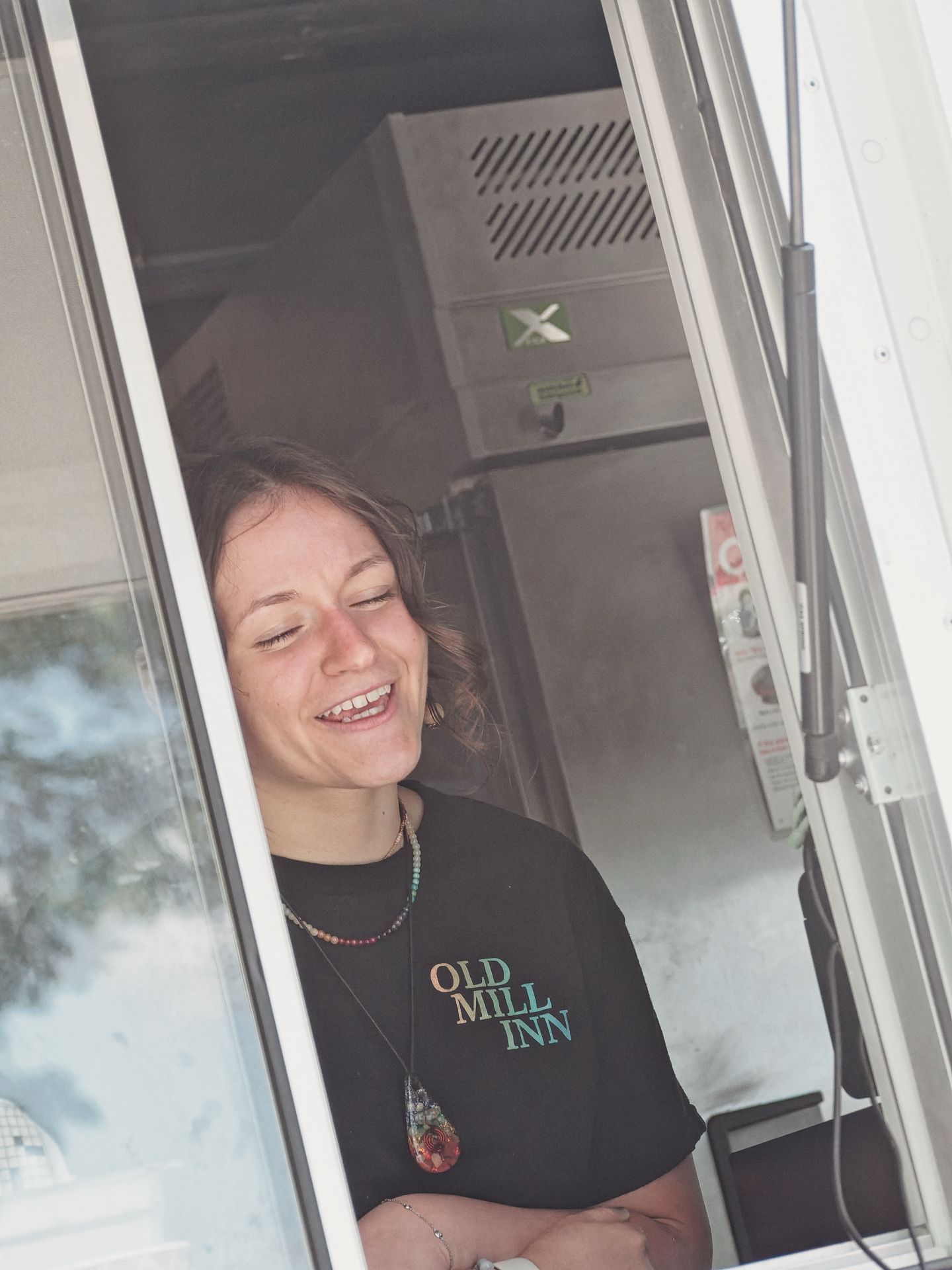 A laughing young woman with windswept hair leans in the service window of a food truck, wearing a black t-shirt reading "OLD MILL INN" and layered necklaces including a large resin pendant. Kitchen equipment and a green safety sign are visible in the dim interior behind her.