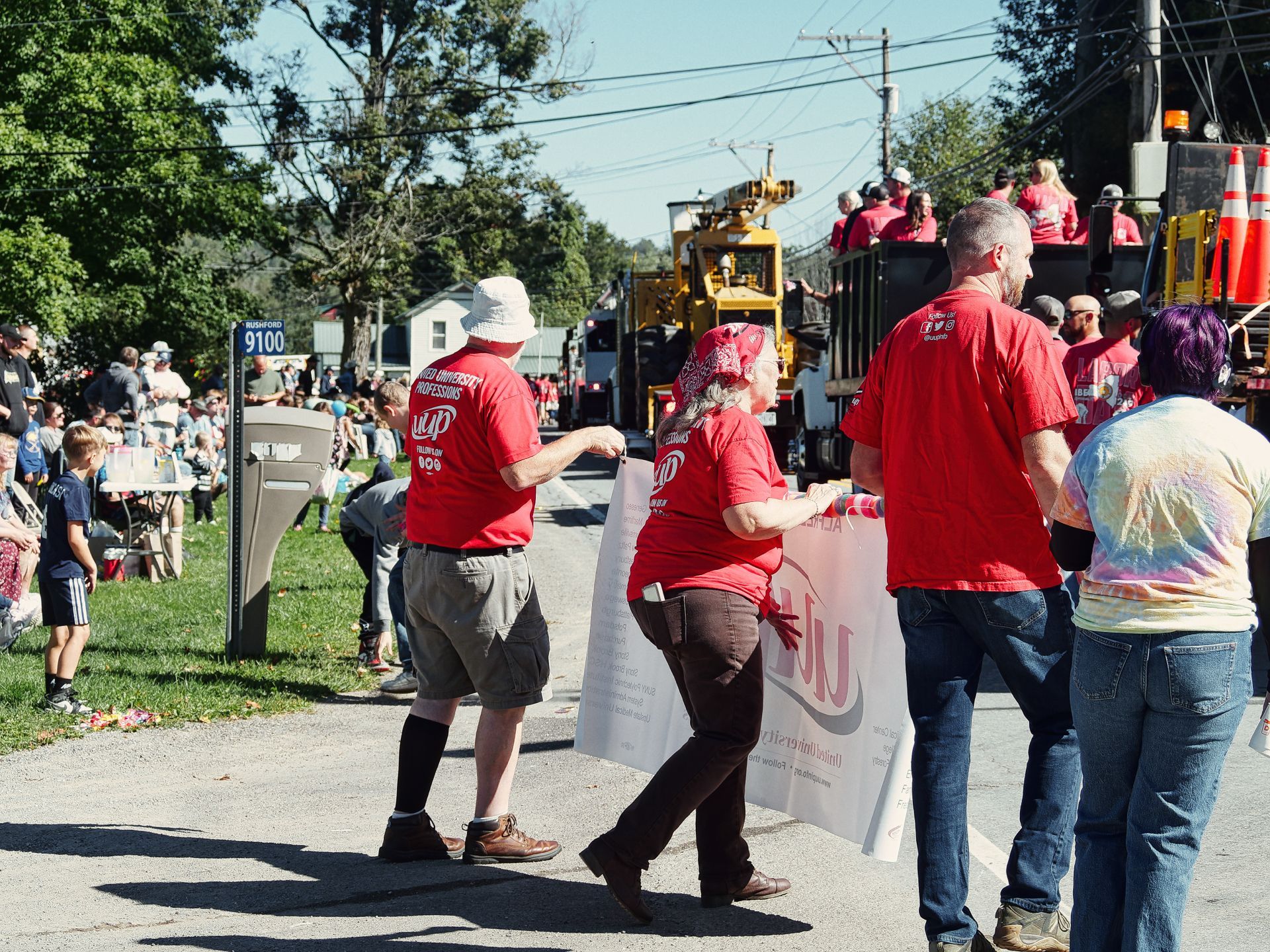 Parade marchers in matching red "IUP University Professions" shirts carry a partially visible banner along a sunny street marked with a "Rushford 9100" sign, while a crowd lines the grass to the left and a yellow construction vehicle and more red-shirted participants ride on a float in the background. A person in a tie-dye shirt walks at the far right, and green trees frame the blue sky above.