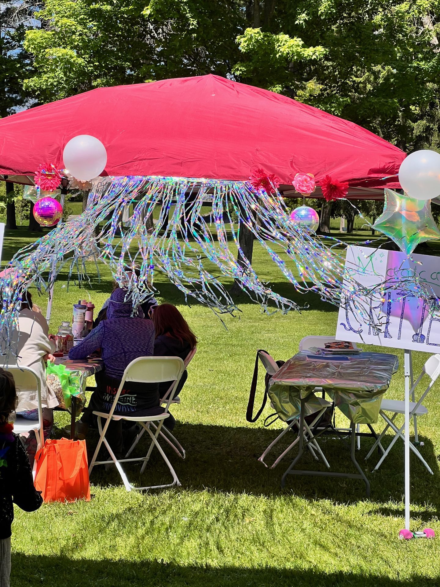 Outdoor vendor or activity booth beneath a large red canopy tent draped with cascading iridescent tinsel fringe, white balloons, pink flower decorations, and small disco balls. Two people sit at a folding table in the foreground, with an orange tote bag on the grass nearby, while a second table covered in holographic silver fabric and a hand-drawn sign on an easel sit to the right — all set on bright green grass with sun-dappled trees in the background.