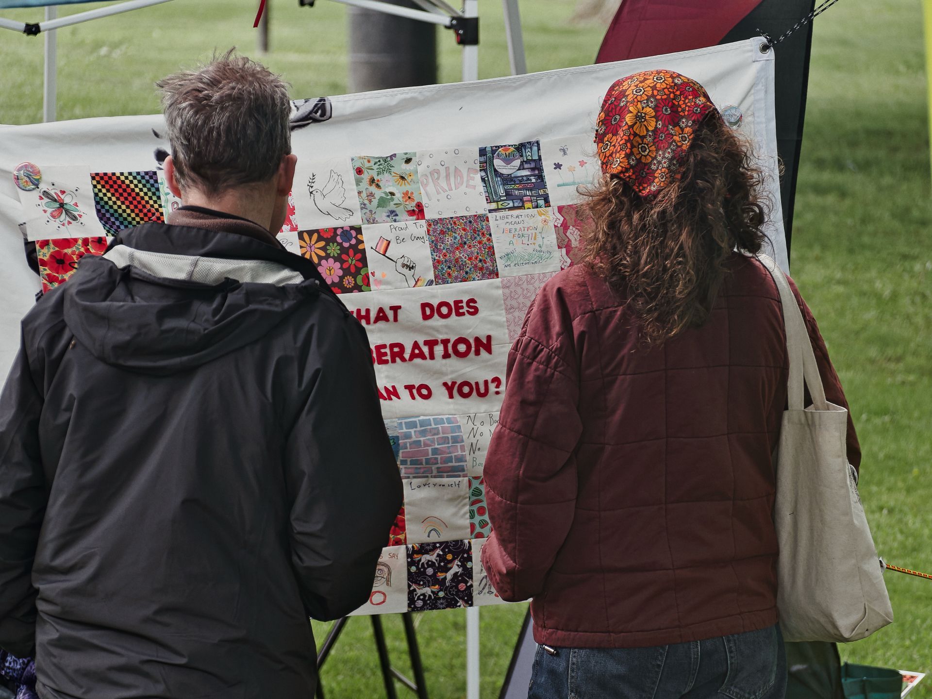 Two people viewed from behind stand at an outdoor event, studying a colorful patchwork banner filled with illustrations, Pride imagery, and the partially visible text "WHAT DOES LIBERATION MEAN TO YOU?" — the person on the left in a dark jacket, the person on the right in a burgundy quilted jacket with a floral headscarf and canvas tote bag. Green grass and event canopy frames are visible in the blurred background.
