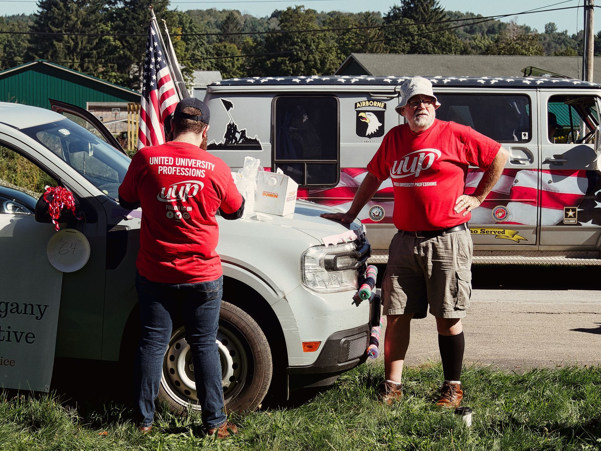 Two people in matching red "UUP – United University Professions" shirts stand on either side of a white SUV at what appears to be a parade or outdoor event, an American flag mounted to the vehicle between them. Behind them, a van decorated with American flags, a 101st Airborne decal, and a "Who Served" banner is parked on the road, with tree-covered hills visible in the background.