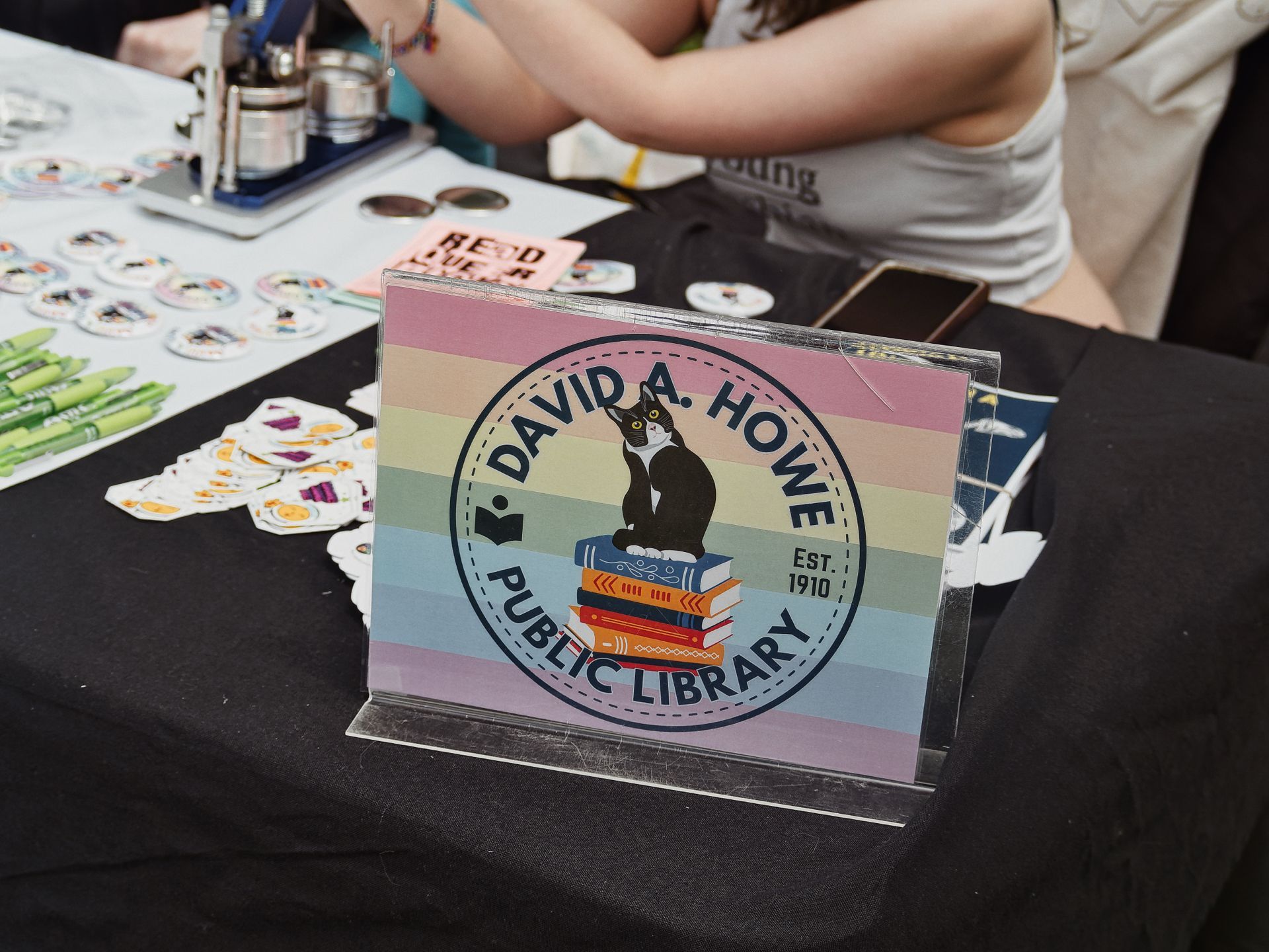 A clear acrylic sign holder on a black-draped table displaying the "David A. Howe Public Library · Est. 1910" logo — a tuxedo cat perched on a stack of colorful books inside a circular badge, set against a rainbow-striped background. Scattered stickers, buttons, green markers, and a pink sign reading "READ" are visible to the left, with a vendor and additional merchandise blurred in the background.