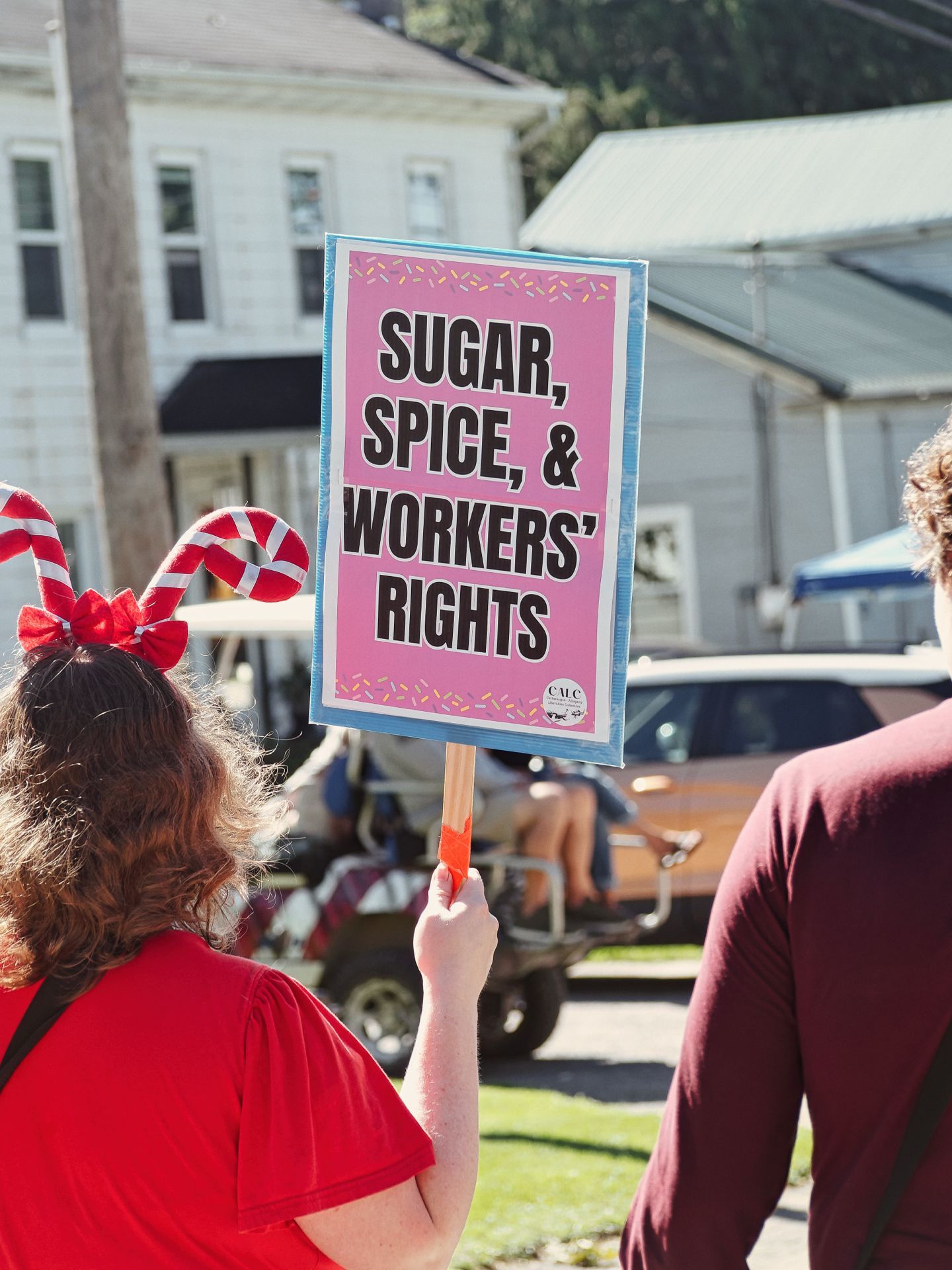Person in a red shirt and candy cane headband holding up a pink protest sign reading "SUGAR, SPICE, & WORKERS' RIGHTS," decorated with colorful sprinkles along the border, with a second person in a maroon shirt to the right and white clapboard buildings blurred in the background.