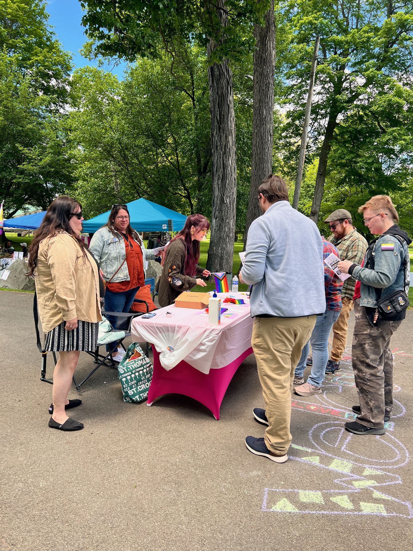 Outdoor Pride event in a park, with a group of people gathered around a table draped in a pink-and-white cloth, topped with a small rainbow flag, pamphlets, and supplies. Chalk drawings decorate the pavement in the foreground, and a person on the right wears a nonbinary pride flag patch on their jacket. A blue canopy tent is visible in the background among lush green trees.