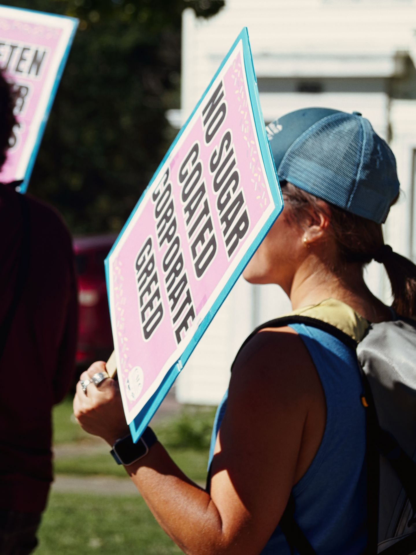Woman in a blue baseball cap and backpack, shot from behind, holding a pink and teal protest sign reading "NO SUGAR COATED CORPORATE GREED." A second partially visible sign to the left reads "TEN" with additional obscured text, held by another person in the foreground, with trees and a white building blurred in the background.