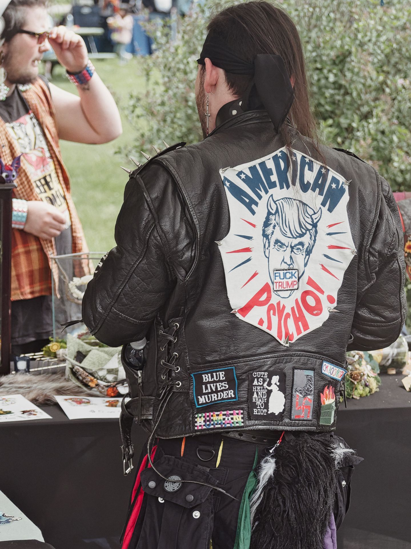 Person seen from behind wearing a heavily customized black leather jacket with metal shoulder spikes and lace-up sides. The back features a large hand-painted patch depicting a devil-horned figure with the text "AMERICAN PSYCHO!" and a "FUCK TRUMP" sticker over the mouth; below it, several sewn patches read "BLUE LIVES MURDER," "#HUMAN," and "CUTE AS HELL AND READY TO RIOT." A second person in a plaid vest stands to the left, partially visible, against a blurred outdoor market backdrop.