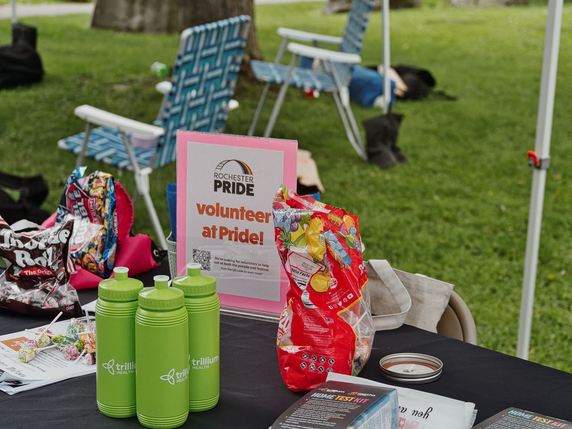 Outdoor table at a Pride event covered in a black cloth, topped with three lime-green "trillium health" water bottles, a pink-backed sign reading "Rochester PRIDE — volunteer at Pride!", a large red bag of mixed candy, lollipops, pamphlets, and "HOME TEST KIT" brochures in the foreground. Two blue-and-white folding lawn chairs and a tree trunk sit blurred on the grass in the background.