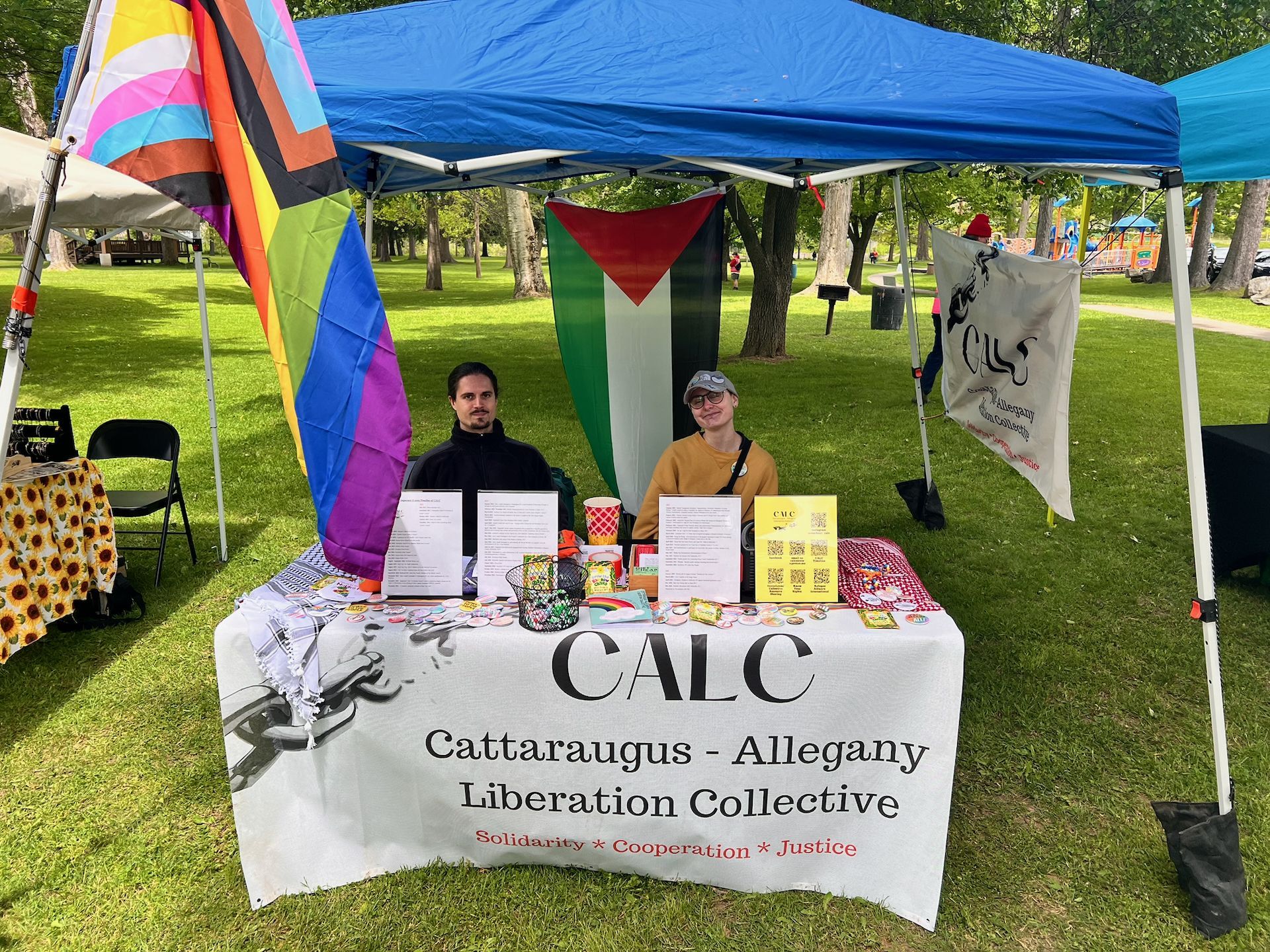 Two people seated behind an outdoor tabling setup under a blue canopy in a grassy park, with a white tablecloth reading "CALC — Cattaraugus - Allegany Liberation Collective — Solidarity * Cooperation * Justice." The table holds printed flyers, buttons, and a yellow QR code sign; behind them hang a Progress Pride flag and a Palestinian flag. A banner on the right canopy pole repeats the CALC logo.