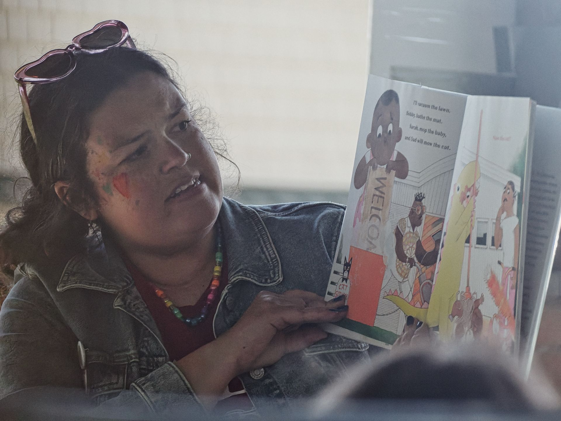 Woman with heart-shaped pink sunglasses pushed up on her head, rainbow bead necklace, and small painted marks on her cheek holds up an open children's book toward an unseen audience. The visible page shows colorful illustrated characters near a "WELCOME" mat with partial text reading "I'll vacuum the lawn, Bobby bathe the mat, Sarah mop the baby, and Dad will mow the cat."