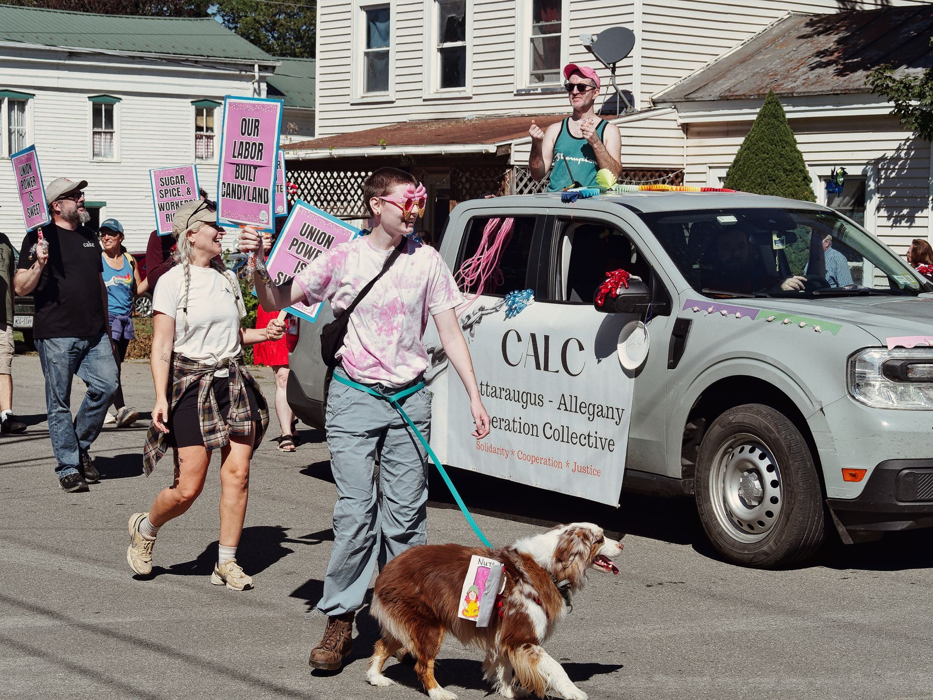 Outdoor labor parade on a sunny street, with marchers carrying pink and teal signs reading "OUR LABOR BUILT CANDYLAND," "UNION POWER IS SWEET," and "SUGAR, SPICE, & WORKERS' RI[GHTS]." In the foreground, a person in a pink tie-dye shirt walks a fluffy brown-and-white dog on a teal leash, a small sign attached to the dog's back. A decorated silver pickup truck to the right displays a banner reading "CALC — Cattaraugus - Allegany Liberation Collective — Solidarity • Cooperation • Justice," with a person standing through the sunroof holding a pink paddle aloft. White clapboard houses line the background.