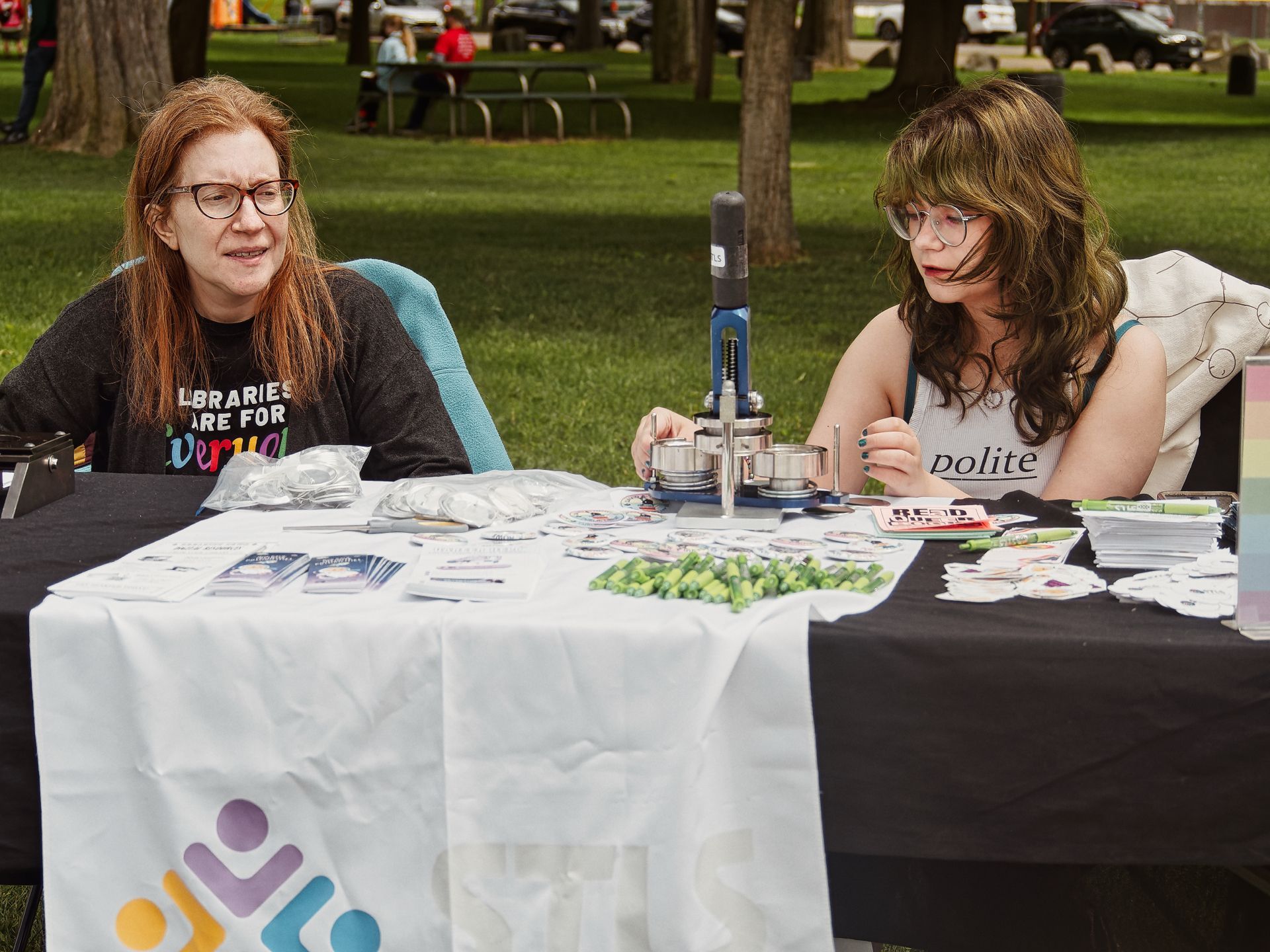 Two women sit behind an outdoor library booth table draped in a white cloth printed with the American Library Association logo. The woman on the left wears a black shirt reading "LIBRARIES ARE FOR EVERYONE" and has long red hair; the woman on the right, with wavy brown hair and round glasses, wears a tank top reading "polite." The table is covered with button-making supplies, small green pencils, pamphlets, stickers, and a metal button press, with a grassy park and blurred trees in the background.