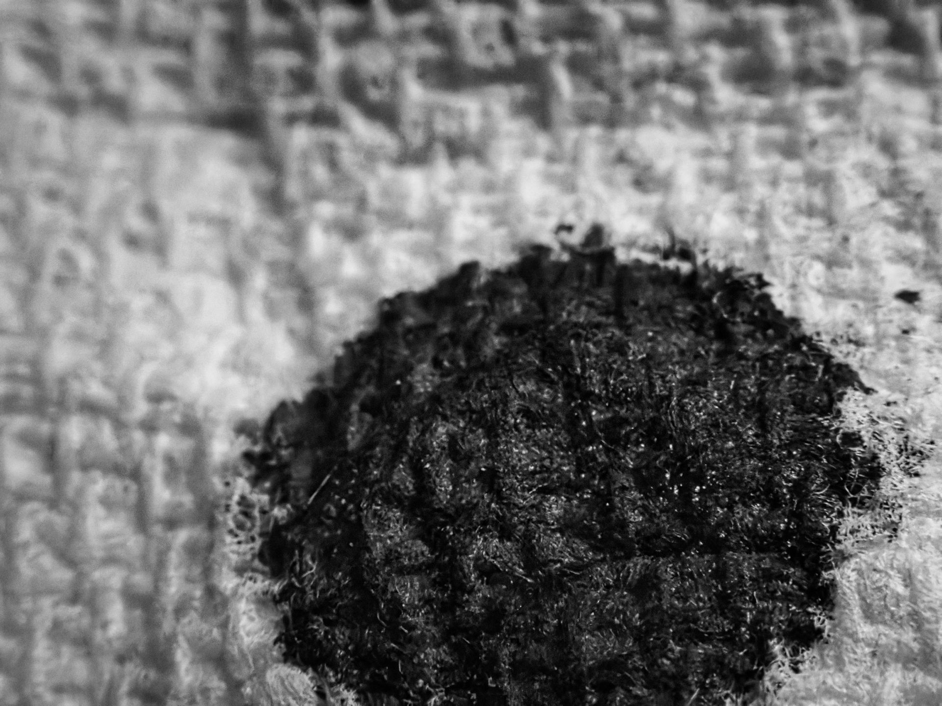 Black-and-white extreme close-up of a dense, rounded ink blot resting on a pale, loosely woven paper towel surface, its fibrous tangle of tendrils nearly planetary in texture against the soft, blurred weave behind it.