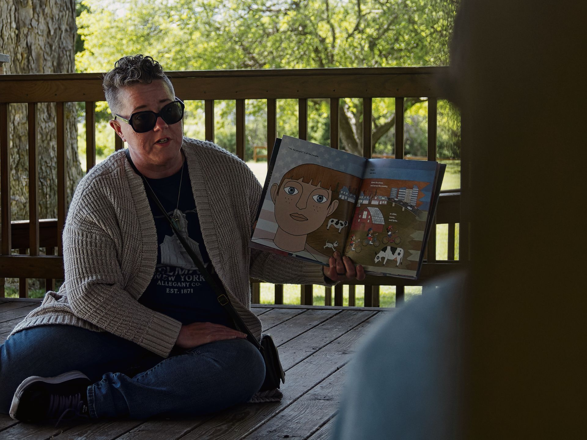 Woman in dark sunglasses and a beige cardigan over a navy shirt reading "ALLEGANY CO. EST. 1871" sits cross-legged on a wooden deck, holding open a children's picture book toward an unseen audience. The open spread shows an illustrated close-up of a freckled, wide-eyed child on the left page and a rural farm scene with cows on the right, with green trees and park grounds visible through the deck railing behind her.