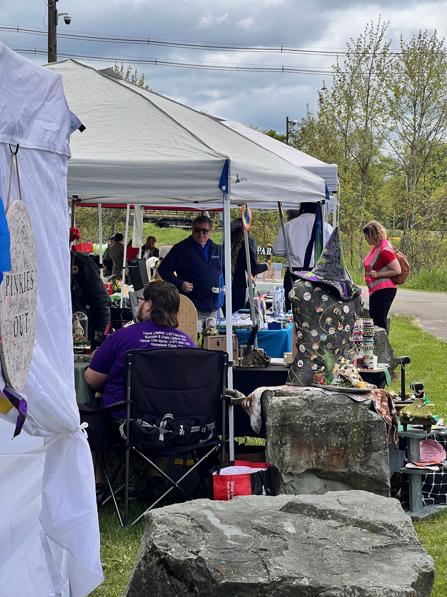 Outdoor craft fair or market under a white pop-up canopy, with vendors and browsing attendees clustered around tables displaying jewelry and small goods. In the foreground, a person in a purple shirt reading "Trans Lifeline: 1-877-..." sits in a folding chair beside large flat rocks used as display surfaces, topped with a dark star-patterned garment and a witch hat. A hand-painted sign on the left reads "PINKIES OUT," and bare-branched trees line the background under a cloudy sky.