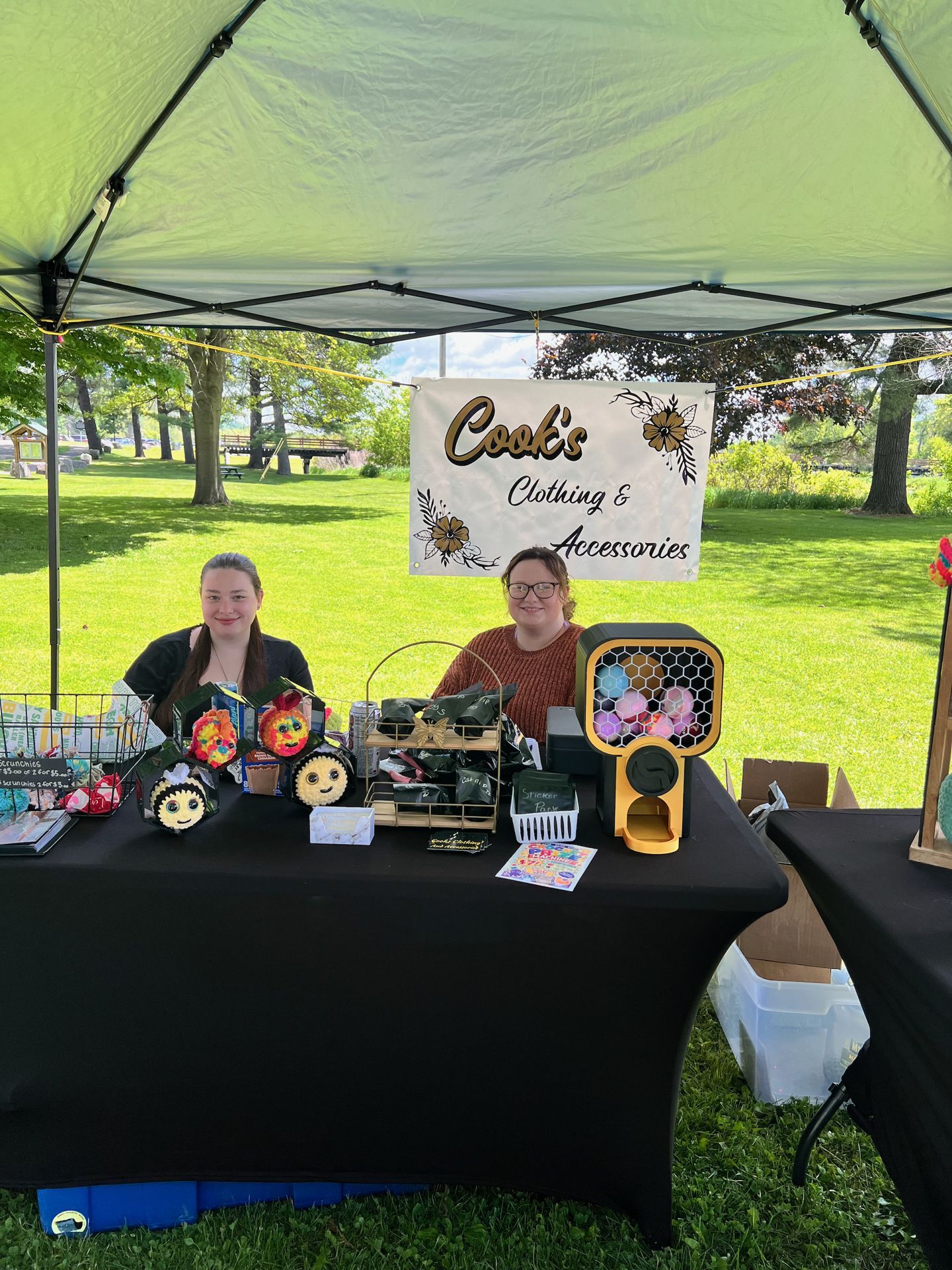 Two women sitting behind a black-draped vendor table at an outdoor craft fair beneath a lime-green pop-up canopy, with a white banner behind them reading "Cook's Clothing & Accessories" in gold and black lettering with floral accents. The table holds colorful handmade items, black-wrapped packages, a gold wire display rack, and a yellow-framed honeycomb fan, with a sunny park and green trees visible in the background.