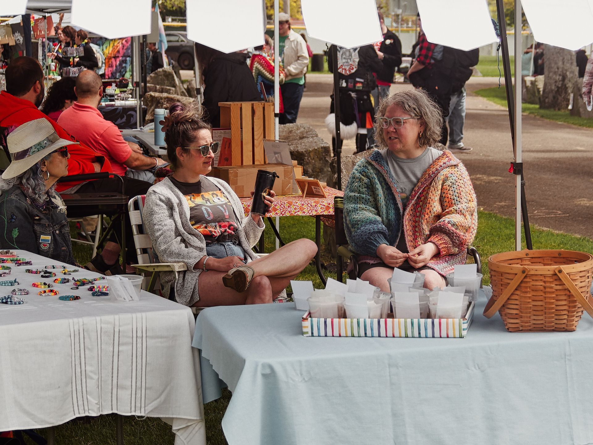 Two women seated behind vendor tables at an outdoor craft fair, mid-conversation — one in a gray cardigan holding a black tumbler, the other in a multicolored crocheted hoodie and glasses. The foreground table holds small packaged goods in a striped rainbow tray and a wicker basket; the left table displays colorful beaded bracelets. A woman in a wide-brim hat sits to the left, and a man in a salmon shirt is visible behind her, with a busy background of white canopy tents, more vendors, and scattered fairgoers on grass and pavement.