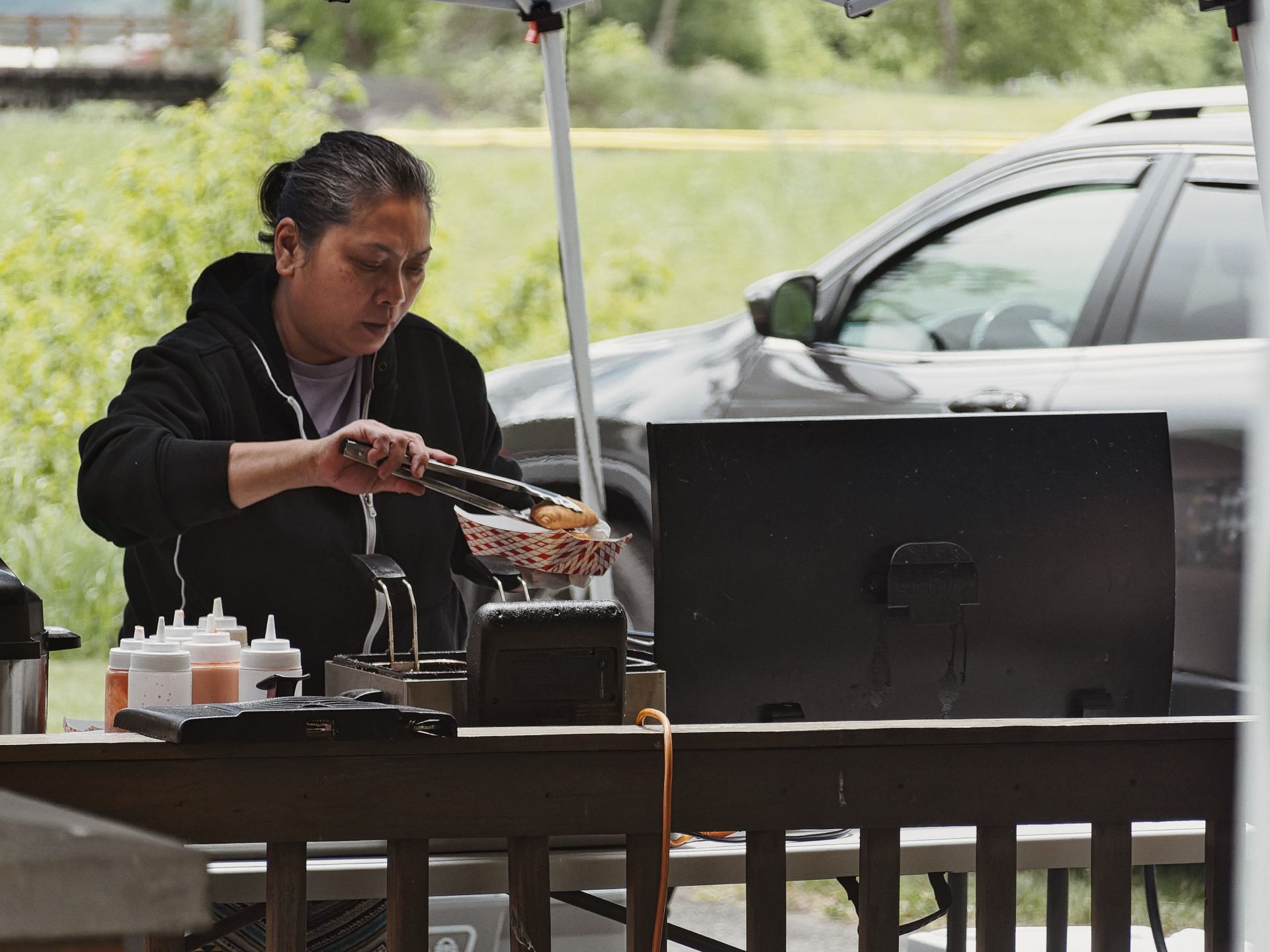 Person with dark hair pulled back, wearing a black hoodie, using tongs to place food into a red-and-white checkered basket at an outdoor food stand under a canopy. A row of squeeze bottles and condiment containers lines the table to the left, beside a large black griddle or smoker; a dark SUV and lush green foliage fill the blurred background.