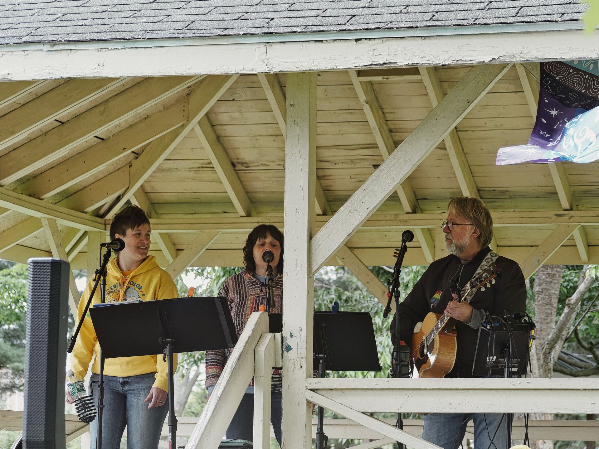 Three performers on a weathered white wooden gazebo stage: a young person in a yellow hoodie smiling at a mic stand on the left, a woman in a patterned top singing into a second mic in the center, and an older man with glasses and long gray hair playing an acoustic guitar on the right. Sheet music stands sit in the foreground, a speaker is visible at the far left, and a purple illustrated banner hangs in the upper right corner.