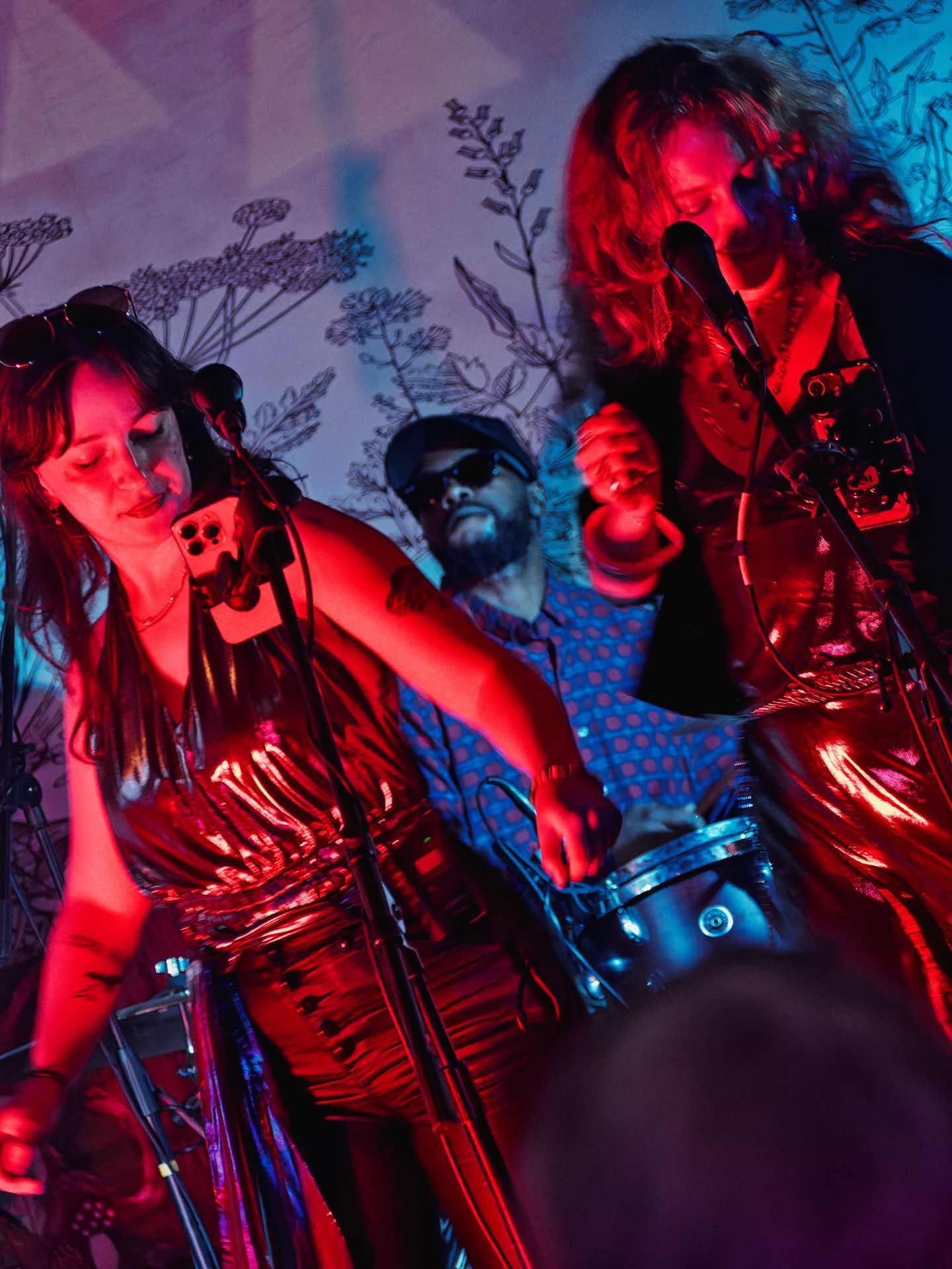 Two performers in matching glossy red jumpsuits lean into microphones under deep red stage lighting, one on the left with eyes closed mid-song, the other on the right with long wavy hair obscuring their face. A bearded drummer in a blue polka-dot shirt and dark sunglasses watches from the background, framed by a wall mural of delicate botanical line drawings.