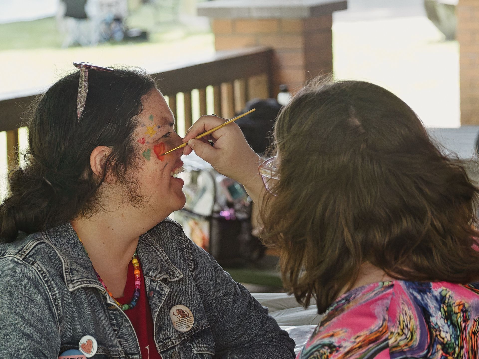 A child in a colorful tie-dye shirt applies face paint to a smiling adult woman in a denim jacket and beaded necklace, using a thin yellow-handled brush to add to the red, orange, and green designs already covering her cheeks. A wooden deck railing and sun-lit lawn are blurred in the background.