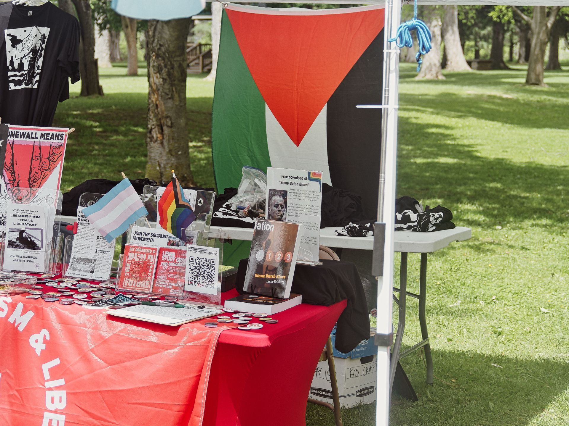 An outdoor tabling setup in a park, with a red-draped table covered in buttons, pamphlets, and small trans pride and rainbow flags. A Palestinian flag hangs as a backdrop alongside a black banner. Visible text includes "STONEWALL MEANS," "LESSONS FROM TRANS LIBERATION," "JOIN THE SOCIALIST MOVEMENT," "Stone Butch Blues" by Leslie Feinberg with a QR code for a free download, and a red pamphlet reading "GET ORGANIZED A PCLU FOR ORGANIZERS." A black t-shirt reading "LIBERATION" hangs to the upper left, and folded black shirts sit on a second table to the right, with tree-lined grass visible in the background.