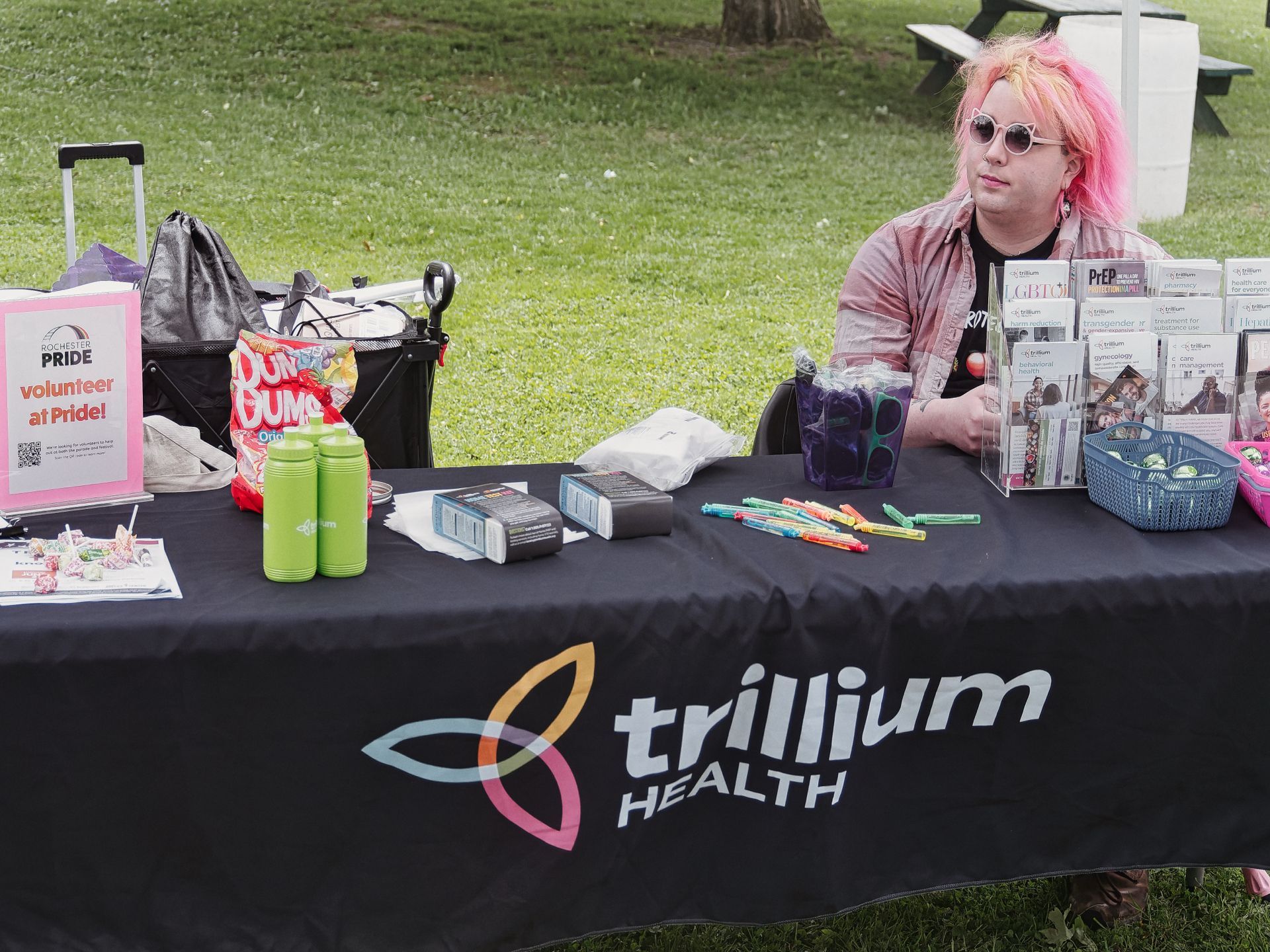 Person with pink hair and round sunglasses sitting behind an outdoor booth table draped in a black "trillium HEALTH" tablecloth. The table holds green tumblers, a bag of Dum Gums, colorful pens, black product boxes, a rack of health brochures including LGBTQ+ and PrEP pamphlets, and a pink sign reading "Rochester PRIDE — volunteer at Pride!" A rolling cart with bags sits to the left, and a grassy park with a picnic table stretches behind them.