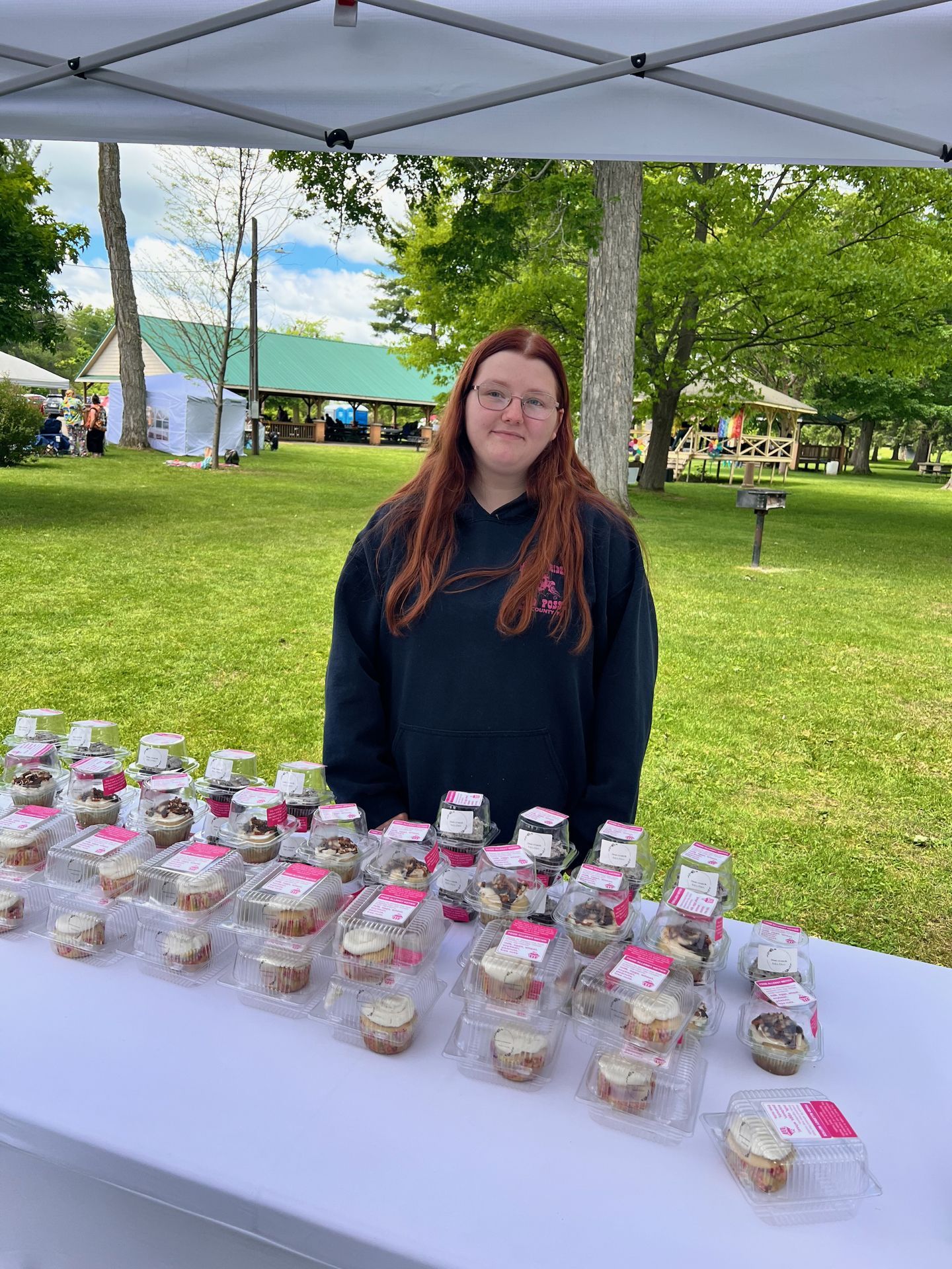 A young woman with long red hair and glasses, wearing a dark navy hoodie, stands behind a white-draped table densely covered with individually packaged cupcakes in clear plastic clamshell containers with pink labels, under a white pop-up canopy at an outdoor market. A green-roofed pavilion and tall trees sit in the sunny park background.
