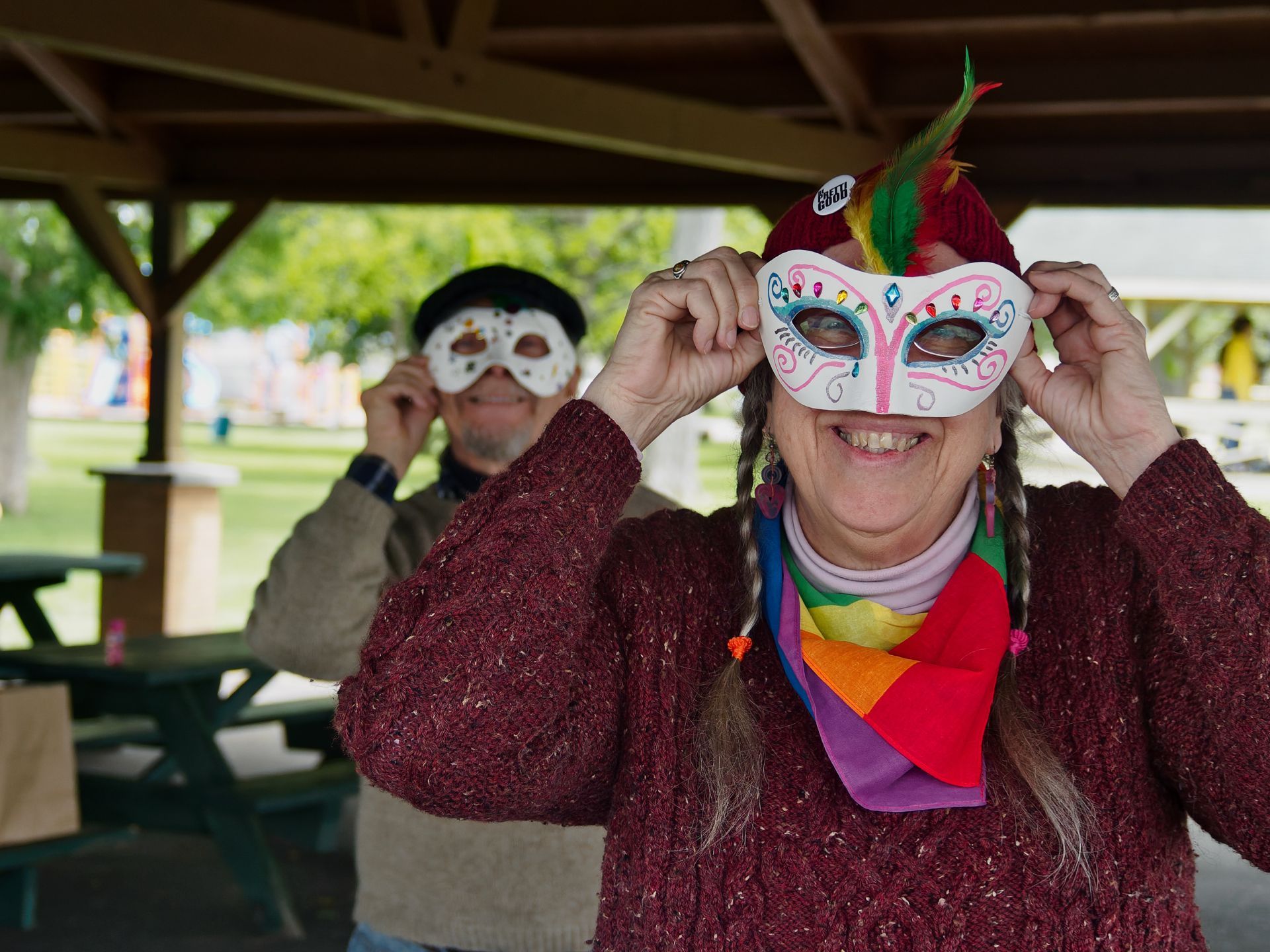 Woman in a burgundy knit sweater, rainbow scarf, and red knit hat adorned with green and red feathers, holding a painted white masquerade mask up to her face with both hands and grinning broadly in the foreground. Behind her under a park pavilion, a man in a flat cap holds a simpler white mask to his face, with green grass and trees blurred softly in the background.