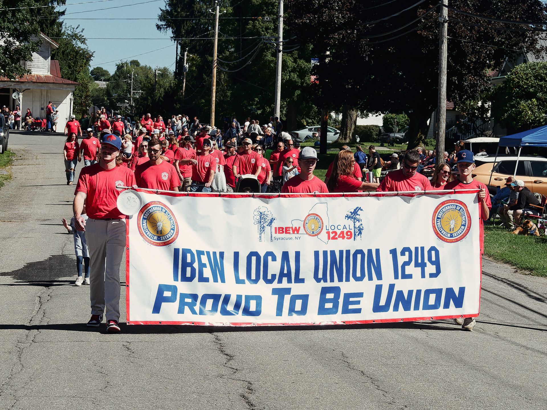 A large group of people in matching red T-shirts march down a residential street carrying a white banner reading "IBEW LOCAL UNION 1249 — Proud To Be Union," with the IBEW Local 1249 Syracuse, NY logo centered above the text. Spectators line the right sidewalk beneath trees, with a blue tent and a vintage gold car visible in the background right.