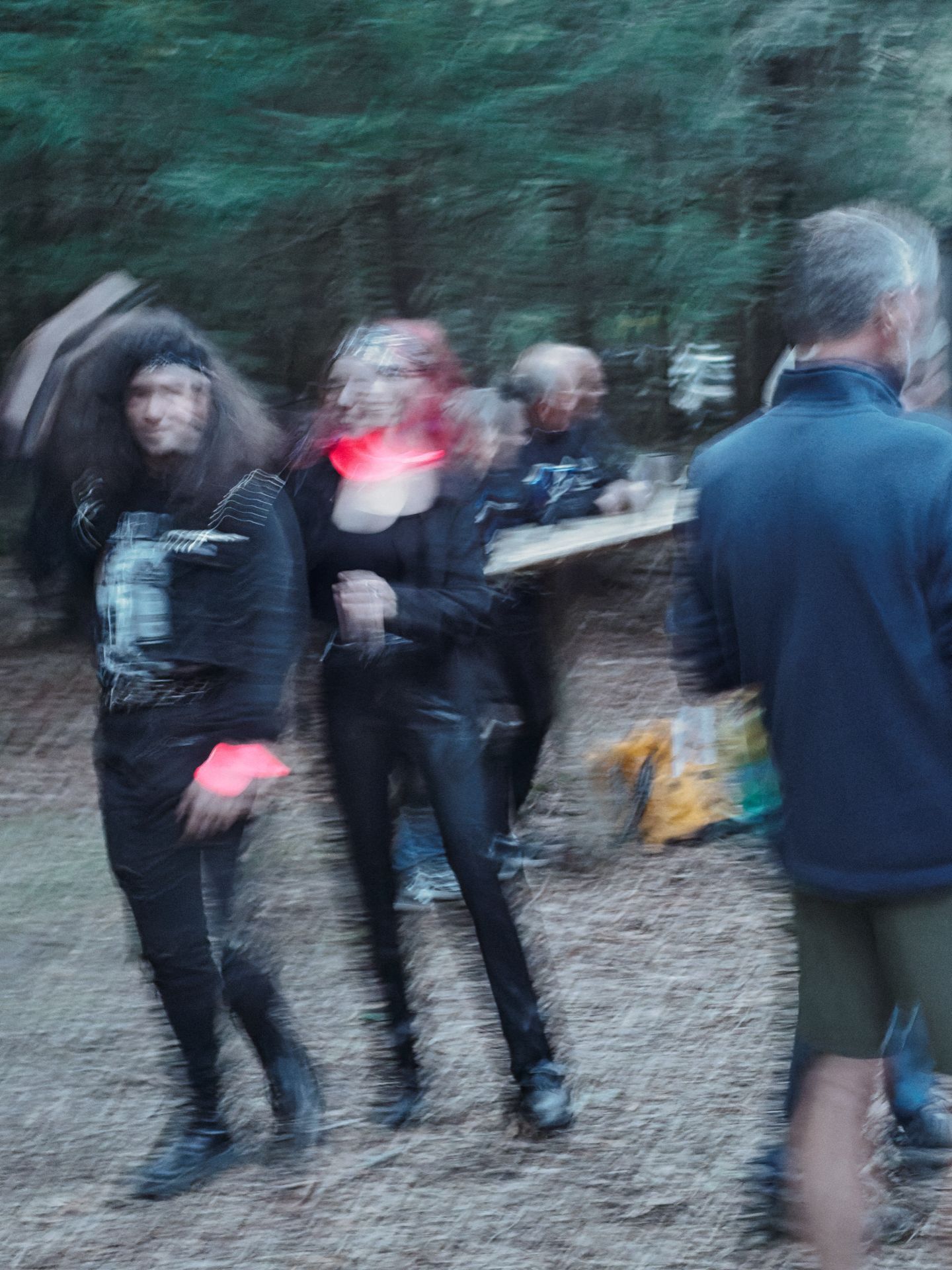 Motion-blurred group of people gathered outdoors on a pine-needle-covered forest floor, with dark trees filling the background — two figures in all-black clothing with flashes of red move in the foreground left, while a man in a blue jacket stands sharply to the right, and a folding table with cups is visible mid-ground.