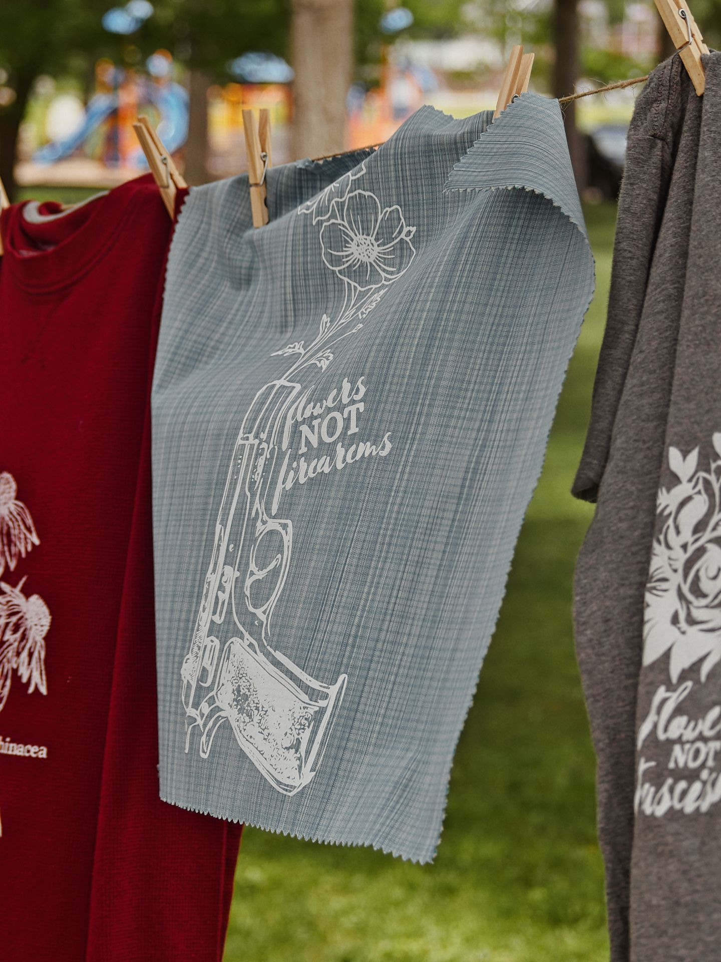 Three items clipped to an outdoor clothesline in a park, with a playground blurred softly in the background. The center piece is a pale blue fabric panel printed with a handgun blooming flowers from its barrel and the text "flowers NOT firearms"; flanking it are a crimson t-shirt printed with "echinacea" and a floral design, and a charcoal t-shirt partially reading "flowers NOT firearms" as well.