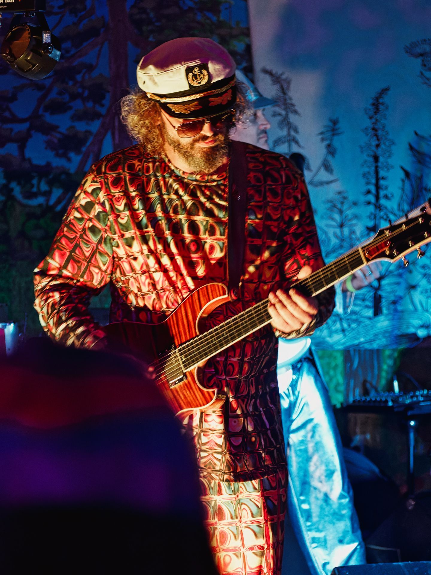 Bearded guitarist in a bold red-and-cream psychedelic patterned jumpsuit, aviator sunglasses, and a white captain's hat with an anchor emblem, playing a warm-toned acoustic-electric guitar on a dimly lit stage bathed in red and blue light, with a painted forest backdrop and a second figure visible and blurred behind him to the right.