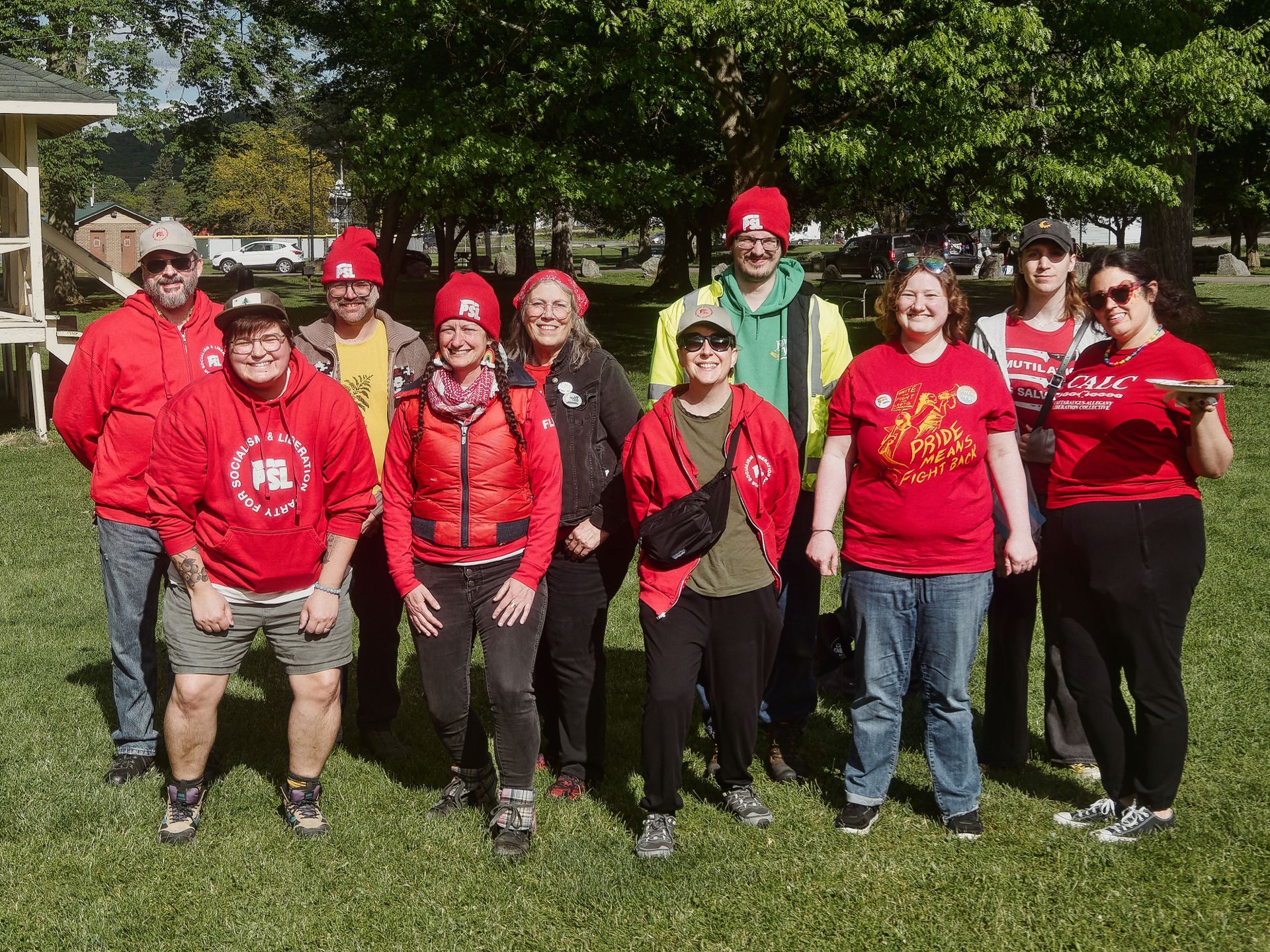 Group of about ten people posing together on a grassy park lawn, most wearing red hoodies, red puffer vests, or red T-shirts, several in red knit beanies. Visible text includes "PARTY FOR SOCIALISM & LIBERATION / PSL" on a hoodie at left, "PRIDE MEANS FIGHT BACK" on a T-shirt center-right, and partial "CALC" text on a shirt far right. One person in the back wears a yellow safety vest and green hoodie, standing out against the otherwise red-clad group. Leafy trees and a building with exterior stairs are visible in the sunlit background.