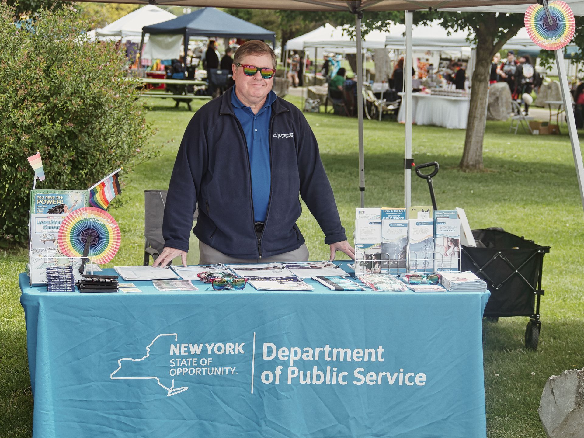 Man in a navy fleece jacket and rainbow-lens sunglasses standing behind an outdoor event table draped in a teal cloth reading "New York State of Opportunity | Department of Public Service." The table holds brochures, pamphlets, pens, and a small rainbow pride fan decoration; a folding wagon sits to the right, and a busy outdoor fair with tents and trees fills the blurred background.