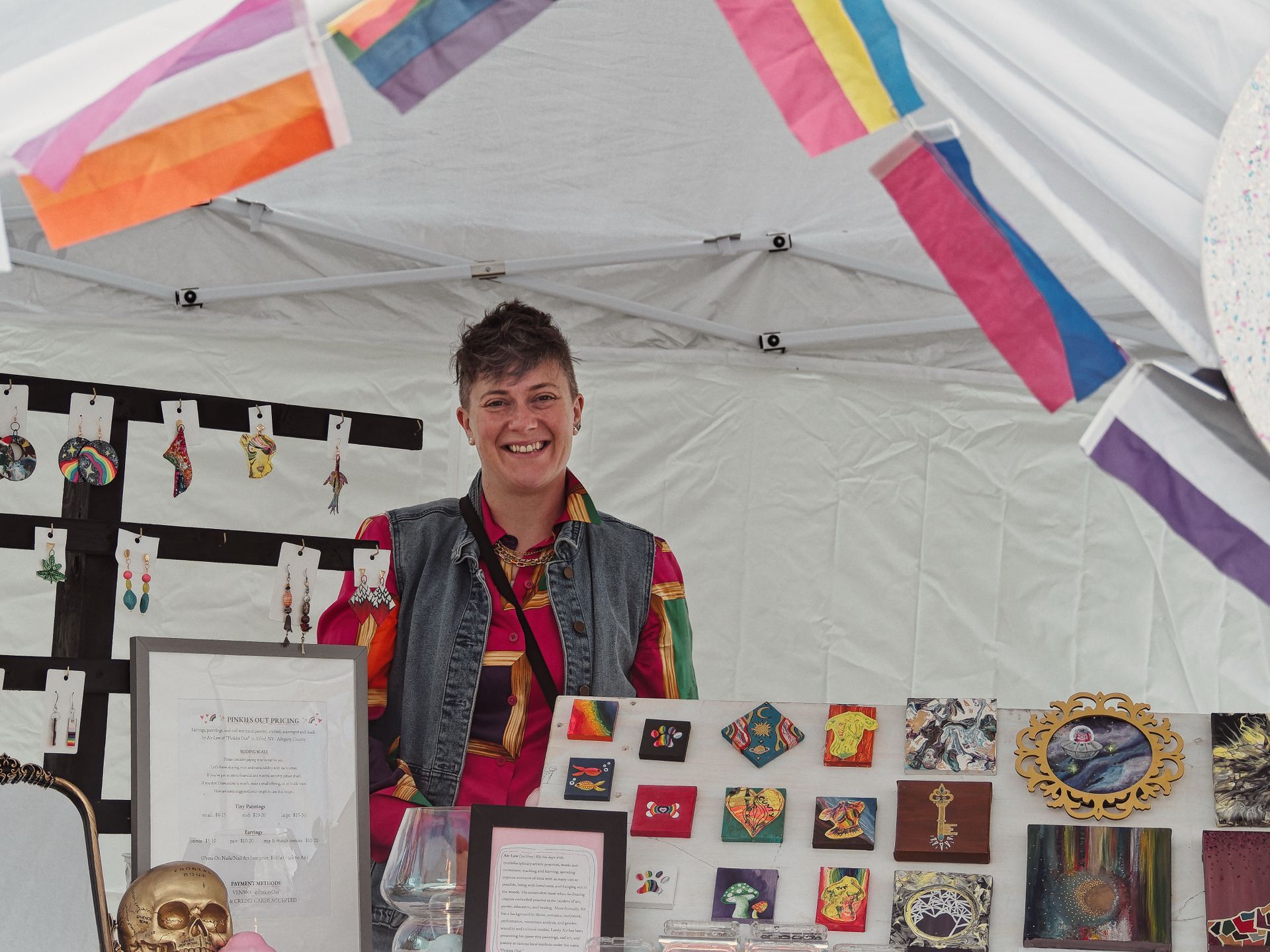 A smiling vendor stands behind a table inside a white pop-up tent, wearing a colorful striped shirt under a denim vest. Pride flags — including bisexual, nonbinary, and intersex flags — hang from the tent frame above, while handmade earrings line a display board to the left, small paintings fill the table and wall to the right, and a gold skull sits in the foreground beside a framed pricing sign reading "PINKIES OUT PRICING."