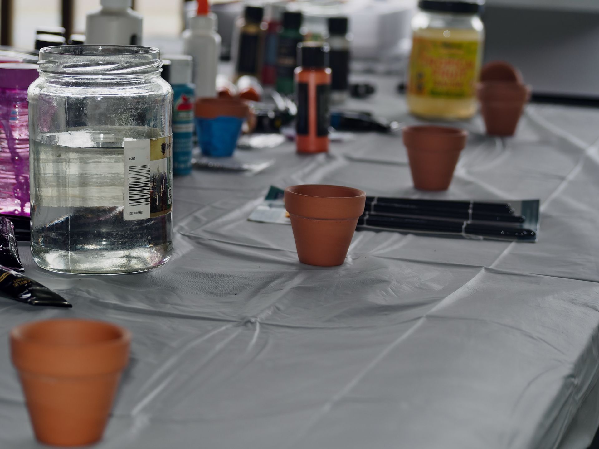 A craft table covered in a gray plastic sheet, scattered with small terracotta pots, a clear glass jar filled with water, paint tubes, and a row of paintbrushes mid-frame. Bottles of acrylic paint, white glue, and a yellow-labeled jar crowd the background, blurred in the shallow depth of field.