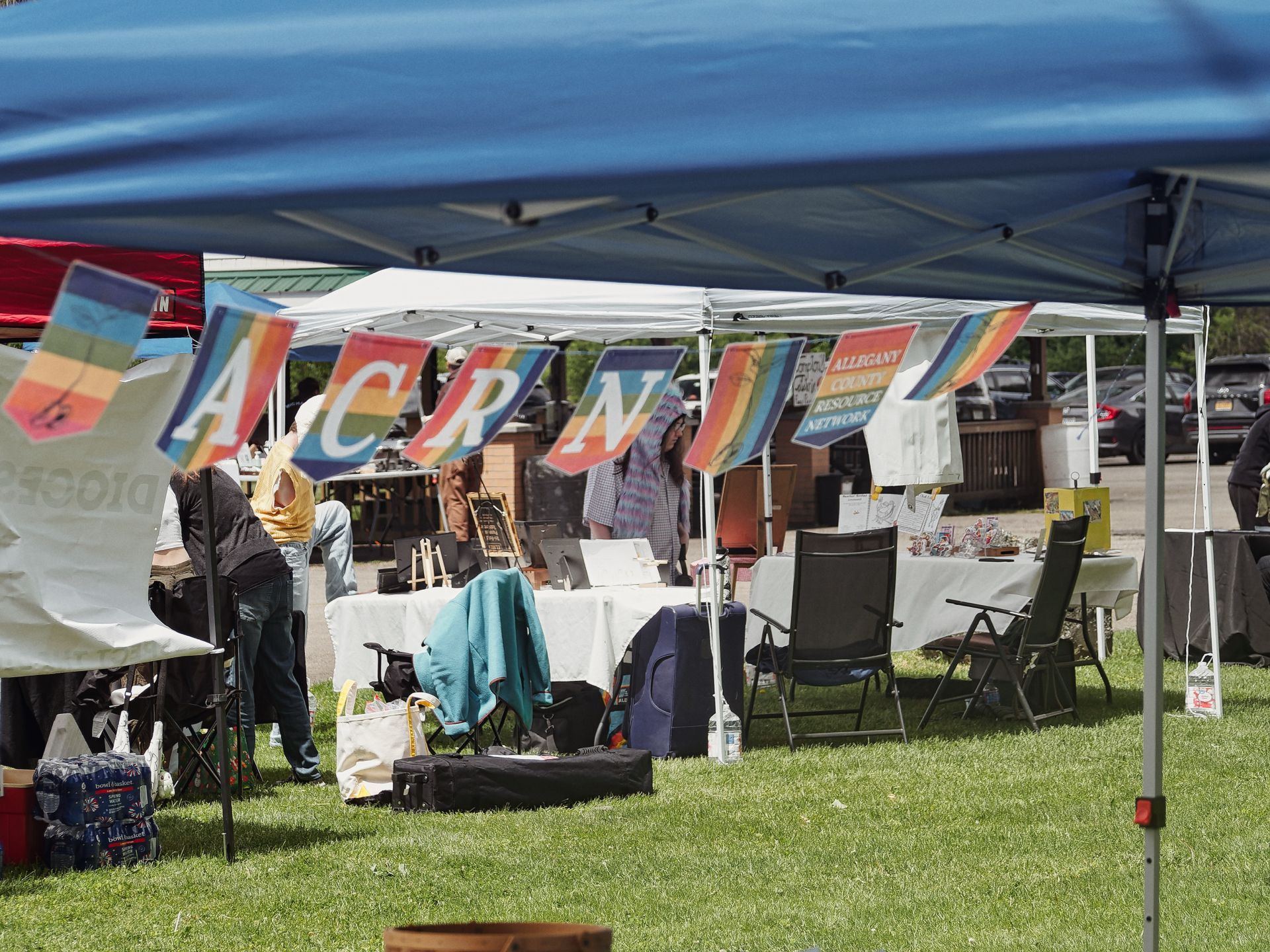 Outdoor community fair on a grassy lawn, shot from beneath a blue canopy tent in the foreground. Rainbow-striped banner flags spelling out "A C R N" hang between white pop-up tents in the middle ground, with a final flag reading "ALLEGANY COUNTY RESOURCE NETWORK" to the right. Several attendees browse vendor tables draped in white cloths, folding chairs scattered around, bags and a case of water bottles on the grass, and a parking lot with parked cars blurred in the background.
