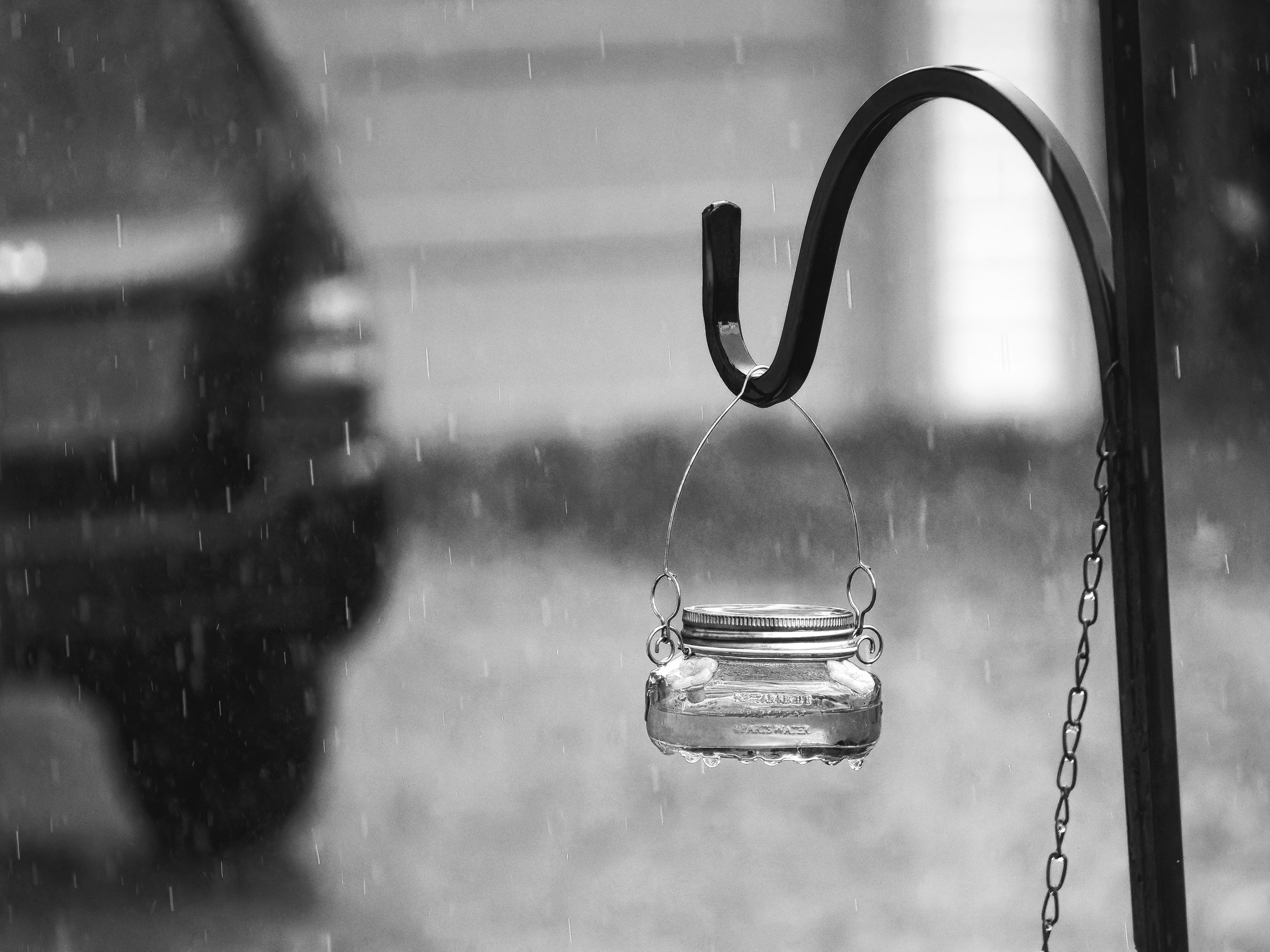 Black-and-white close-up of a small glass mason jar lantern suspended by a wire handle from a curved shepherd's hook, rain streaking visibly through the midground air, with a blurred vehicle and lawn dissolving into shallow depth behind it.