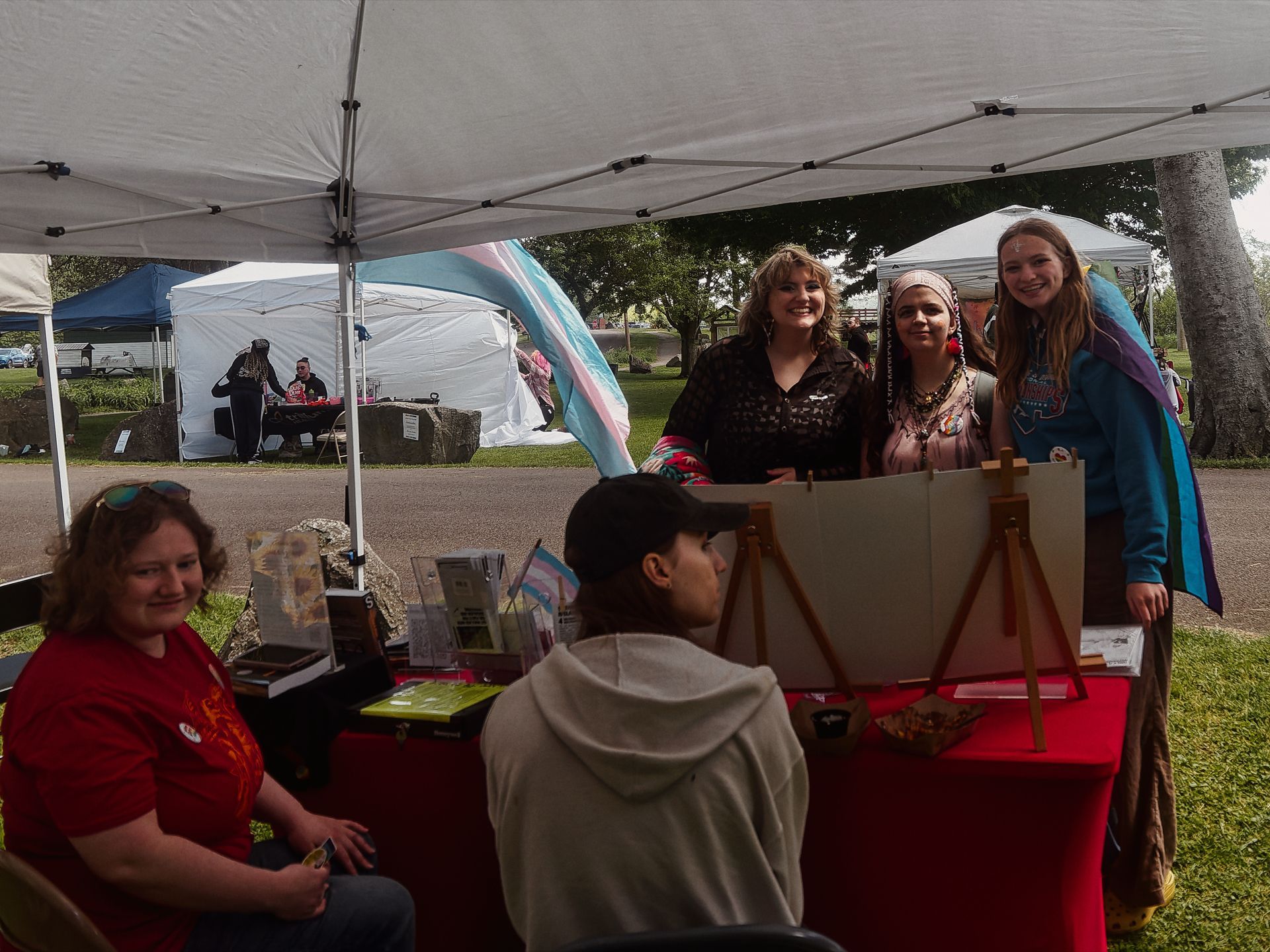 Outdoor vendor booth under a white canopy at what appears to be a community fair, with a red-draped table holding books, small flags, and an artist's easel displaying a blank canvas. Three people stand behind the table posing for a photo — one in a dark patterned blouse, one in layered boho jewelry, and one in a teal hoodie draped with a transgender pride flag in blue, pink, and white. In the foreground, a person in a gray hoodie faces away toward the table, and a woman in a red shirt sits to the left with sunglasses pushed up on her head; additional tents and trees are visible in the background.