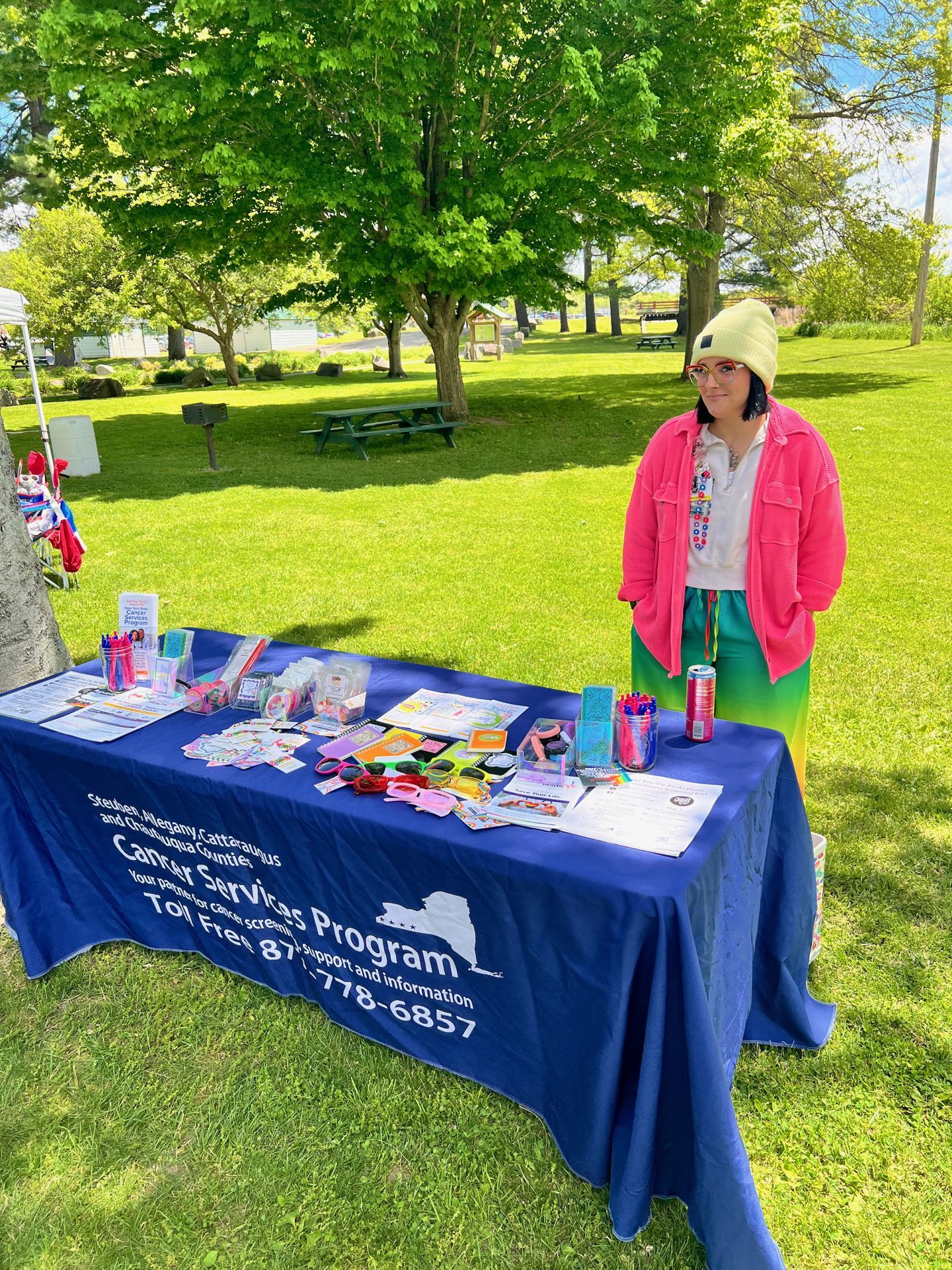 Person in a bright pink jacket, green ombre pants, and a yellow beanie standing beside an outdoor outreach table draped in a royal blue tablecloth reading "Steuben, Allegany, Cattaraugus and Chautauqua Counties Cancer Services Program — Your partner for cancer screening, support and information — Toll Free 877-778-6857." The table is covered in pamphlets, colorful pens, novelty sunglasses, and promotional materials, set on a grass lawn with picnic tables and large trees in the sunny background.
