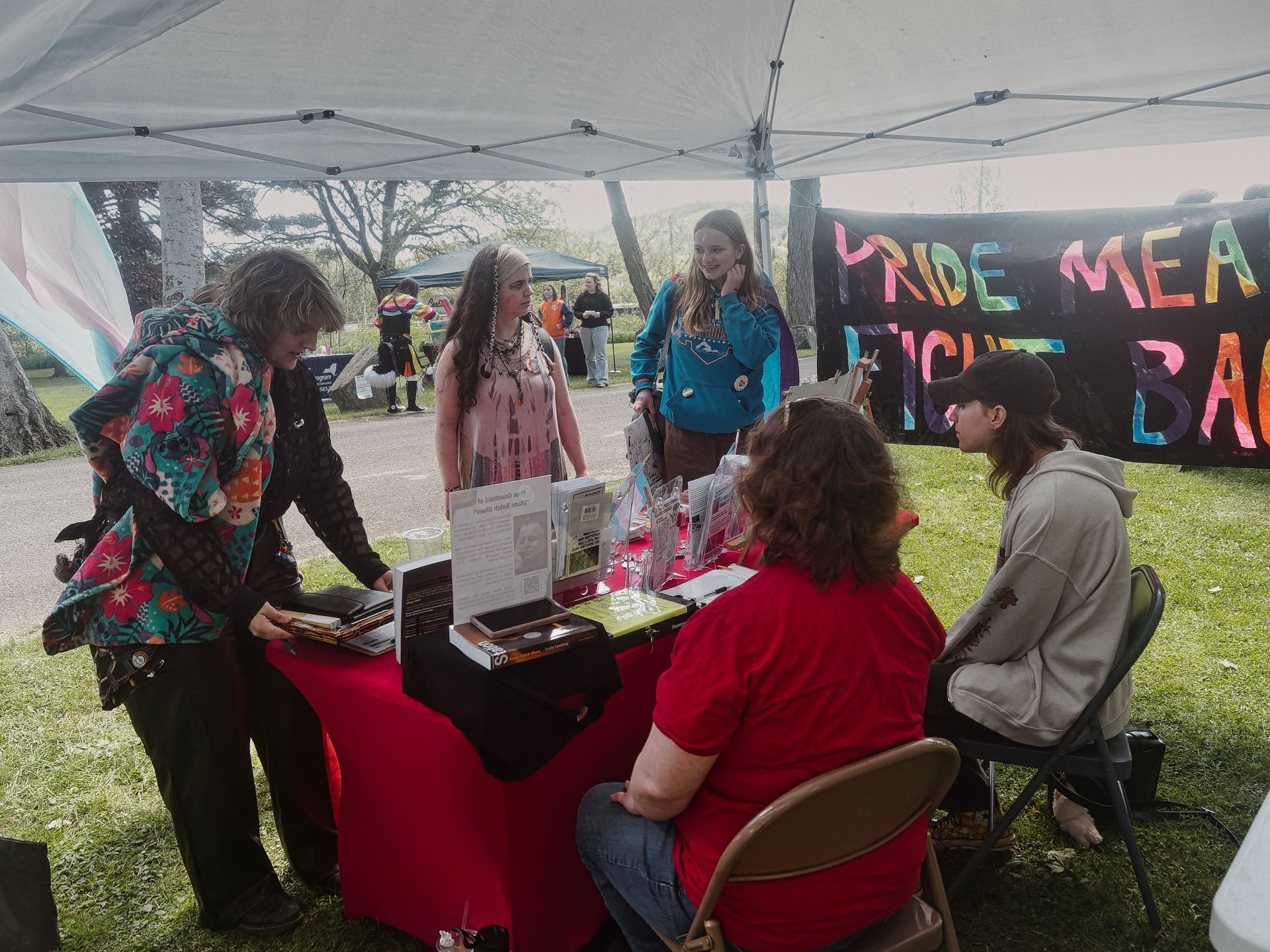 Outdoor Pride event booth under a white pop-up canopy, with a red-draped table covered in pamphlets and display materials. Two people sit behind the table — one in a red shirt, one in a beige hoodie and cap — while three visitors stand across from them, including one in a colorful floral jacket leaning over to look at materials. A hand-painted banner in the background reads "PRIDE MEA[NS] / [F]IGHT BA[CK]" in rainbow letters, and a trans pride flag is partially visible on the left. Trees, grass, and a second green canopy are blurred in the background.